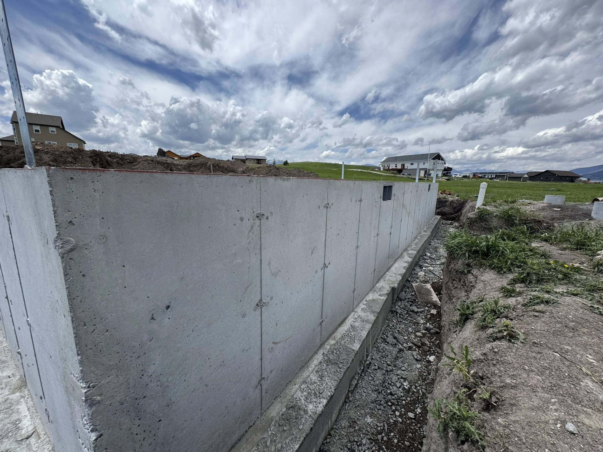 Concrete foundation wall at a construction site with houses and green fields in the background under a partly cloudy sky.