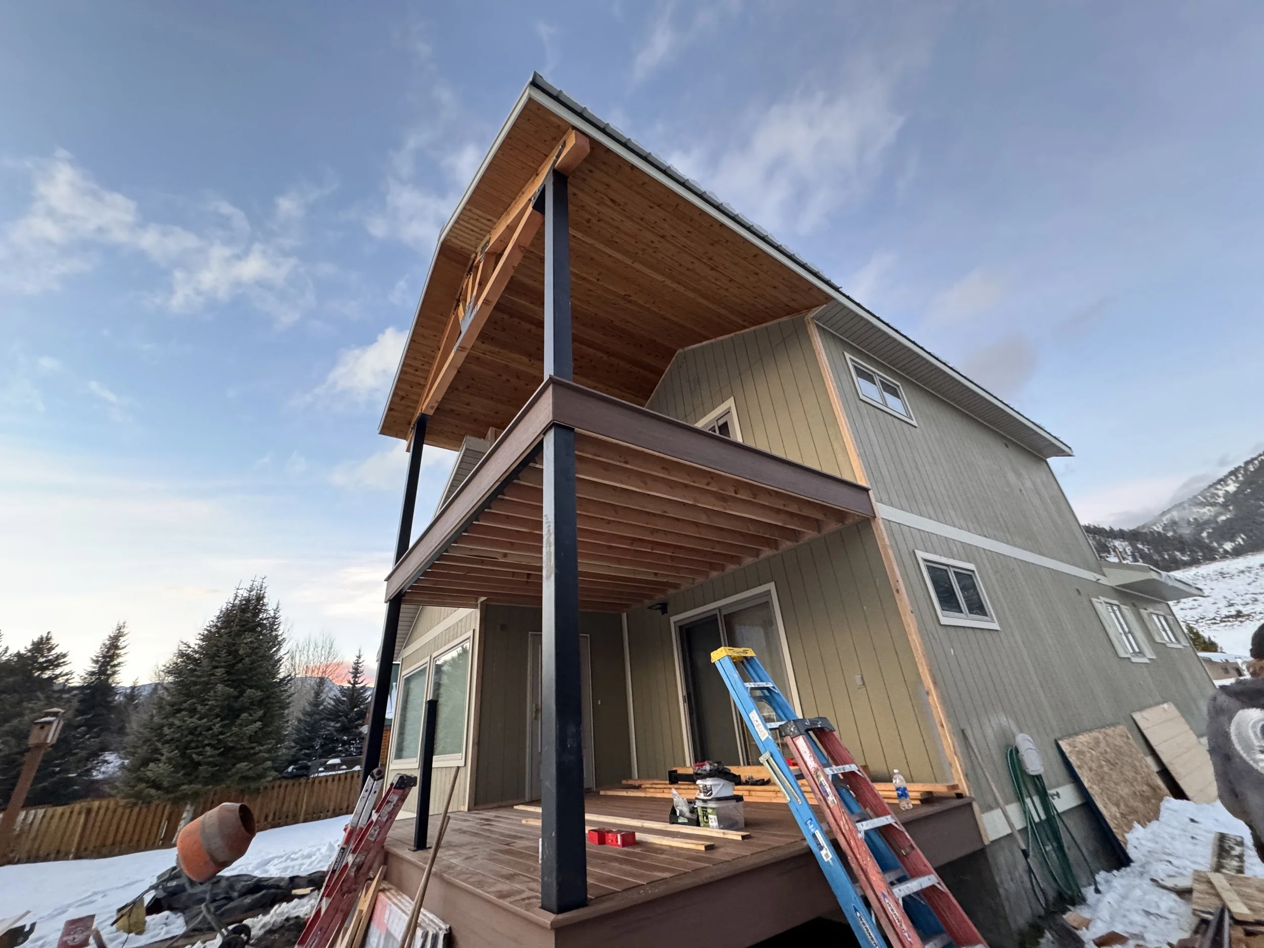 Under construction of a two-story house with a wooden deck and balcony, snow on the ground, and tools and ladders nearby.