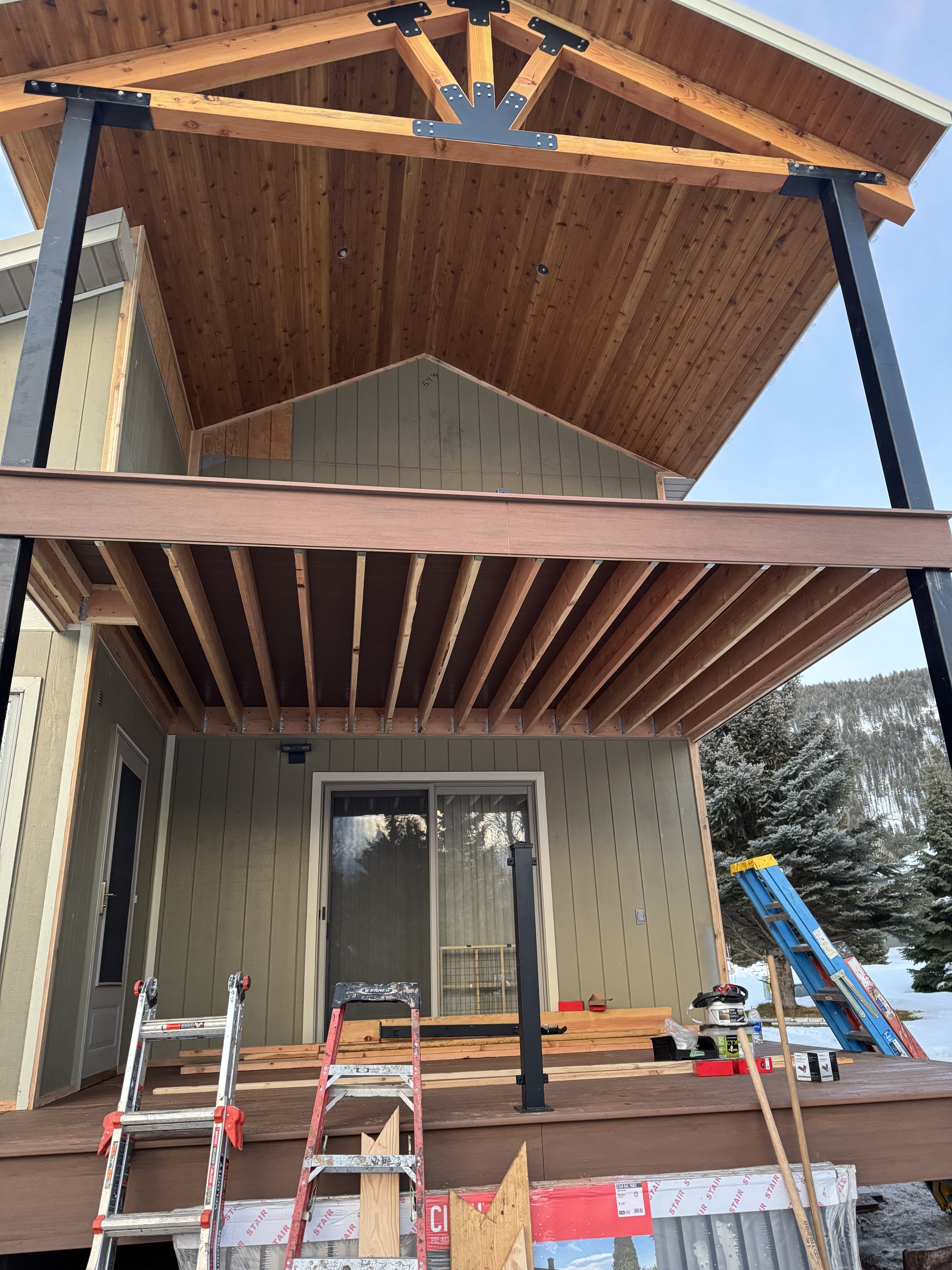 Under construction of two-story house balcony with wooden planks, ladders, and construction tools, mountains with snow and trees in the background.