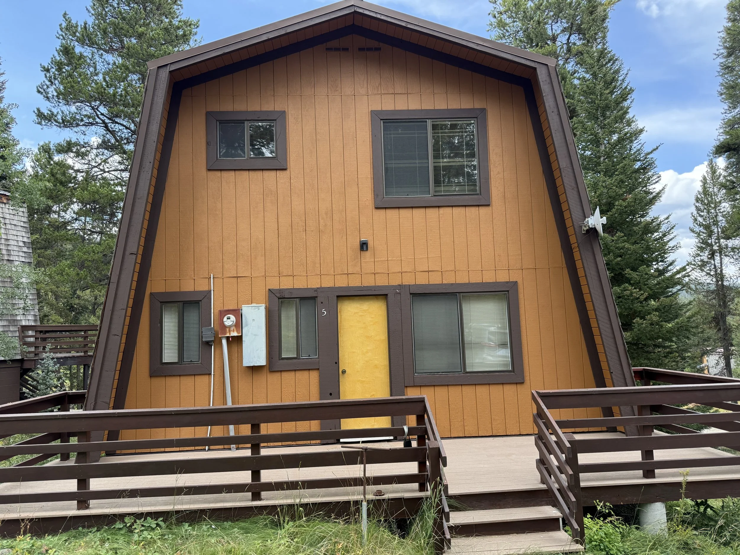 A uniquely designed brown and orange A-frame house with three windows, an orange front door, and a brown wooden deck in a forested area with tall green trees.