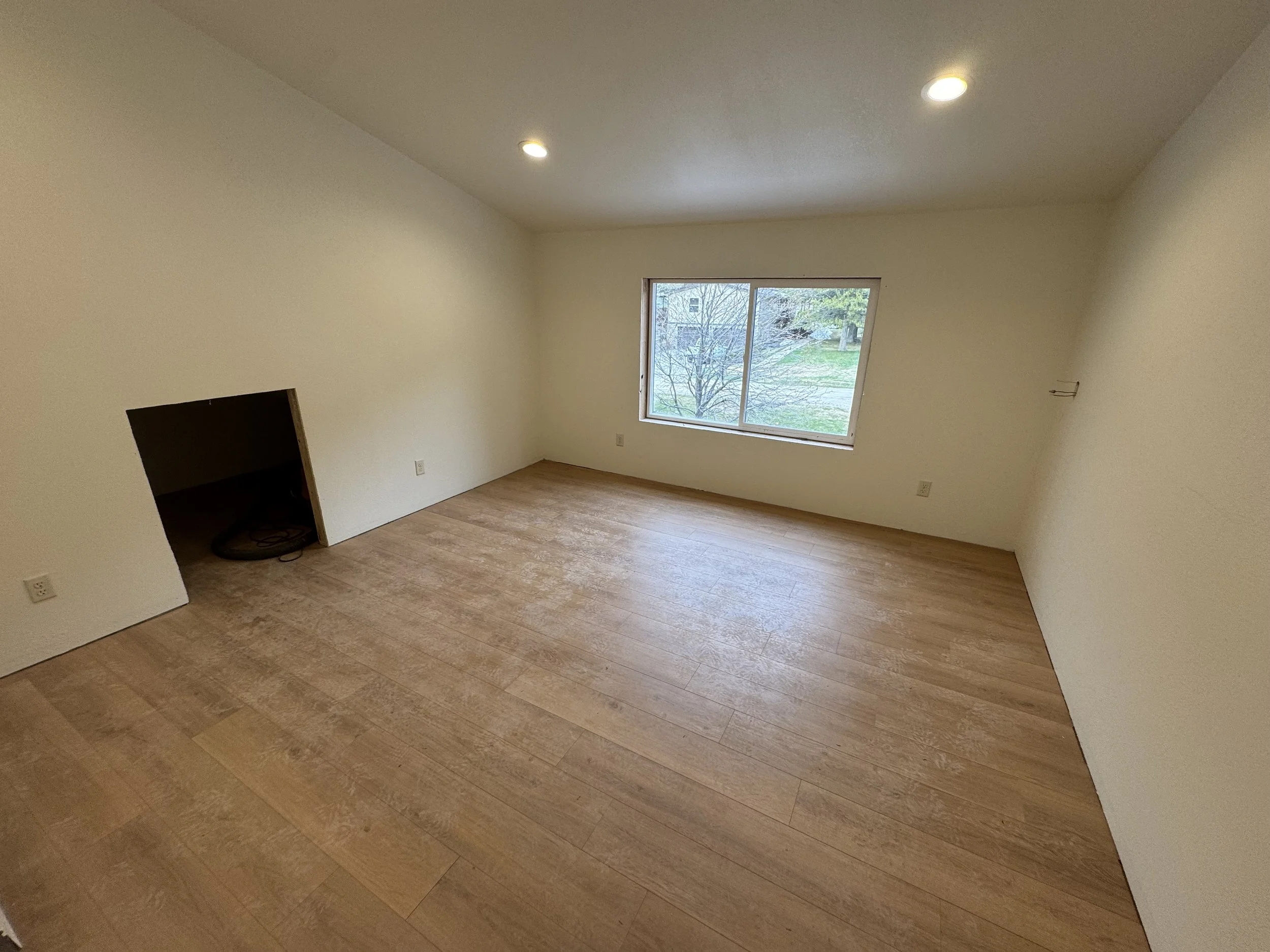 Empty room with hardwood floors, white walls, a window showing trees and grass outside, and a small opening in the wall near the corner.