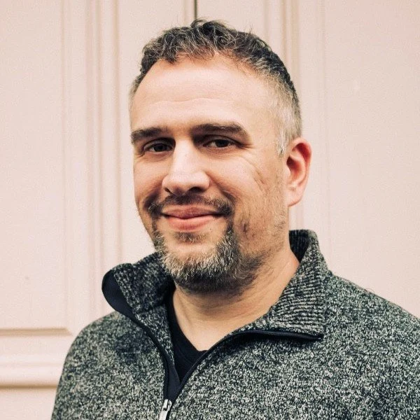 A middle-aged man with short, dark hair and a beard, smiling and wearing a dark gray zip-up sweater, standing in front of a light-colored wooden panel wall.