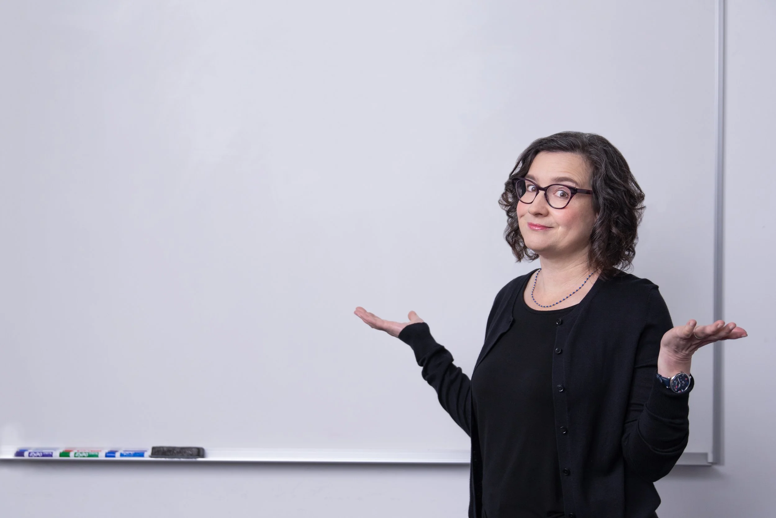 Marianna stands in front of a white board with her arms raised, as if she is asking a question.