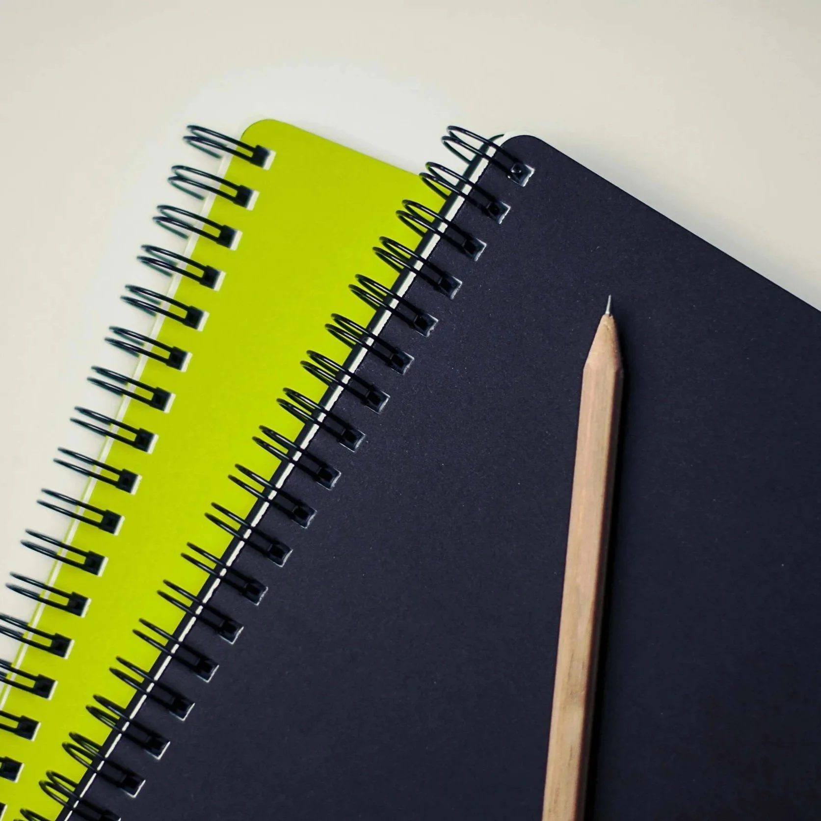 Close-up of three spiral notebooks in yellow, black, and gray colors, with a wooden pencil resting on the black notebook, on a light-colored surface.