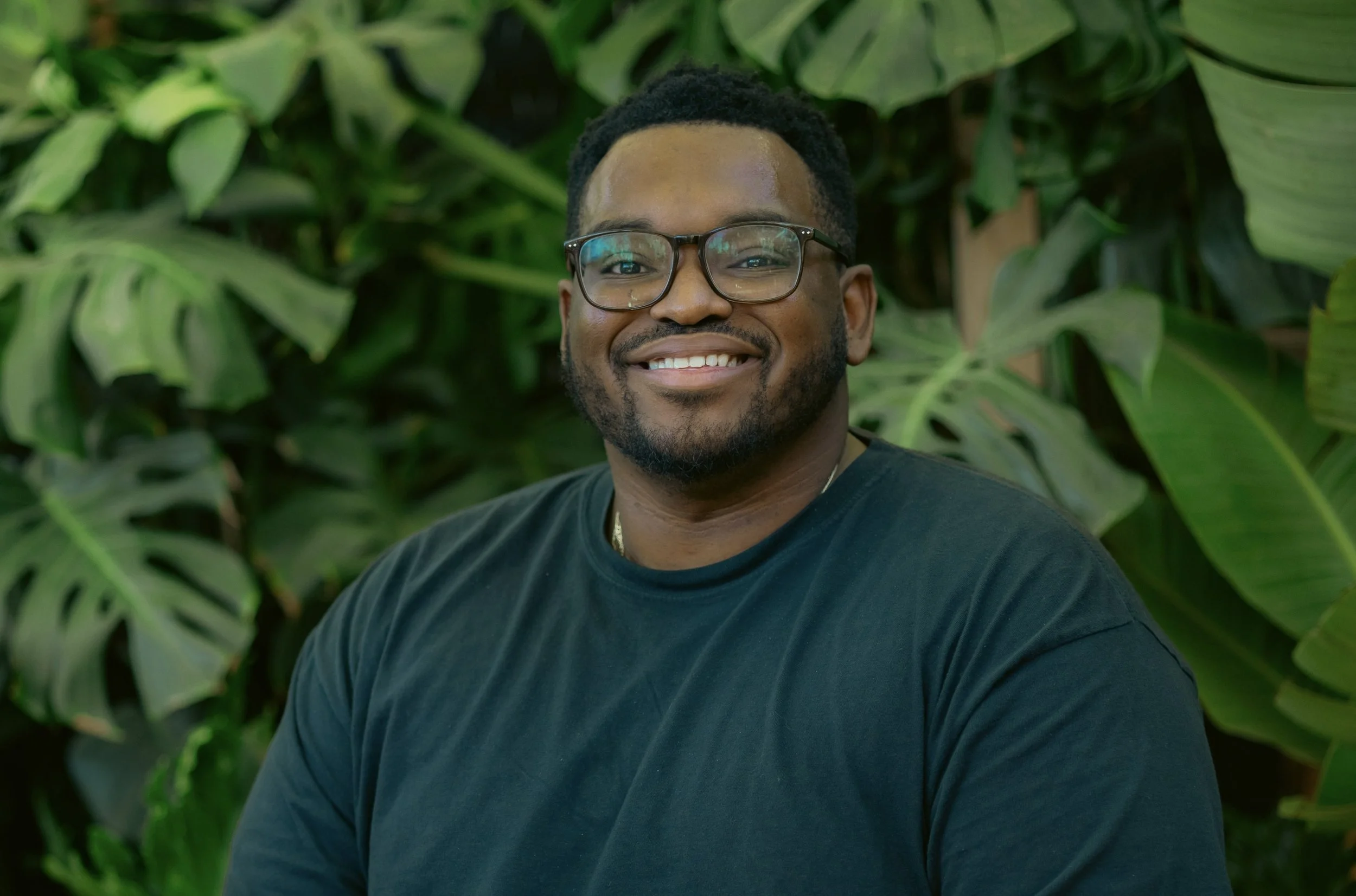 A smiling man with glasses and a beard, wearing a black T-shirt, standing in front of large green leaves.