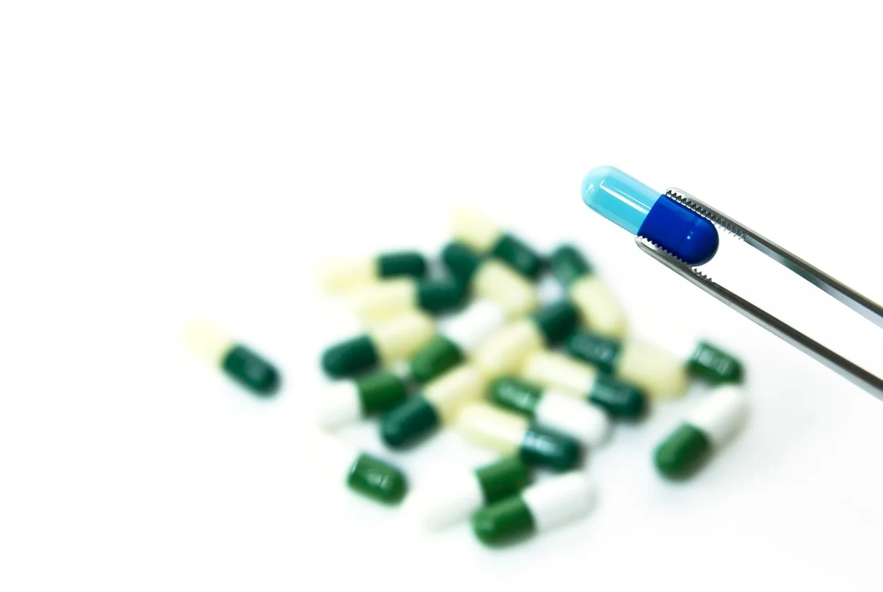 Blue pill being held by surgical tweezers with faded green pills in the background