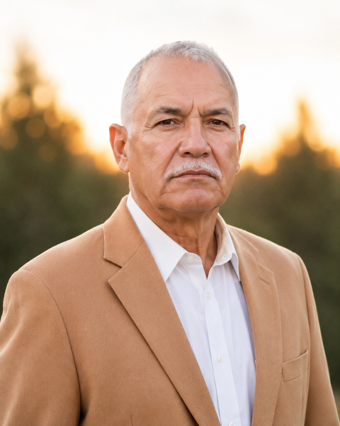 A middle-aged man with short gray hair standing outdoors, wearing a tan shirt with a collar, in front of green trees.