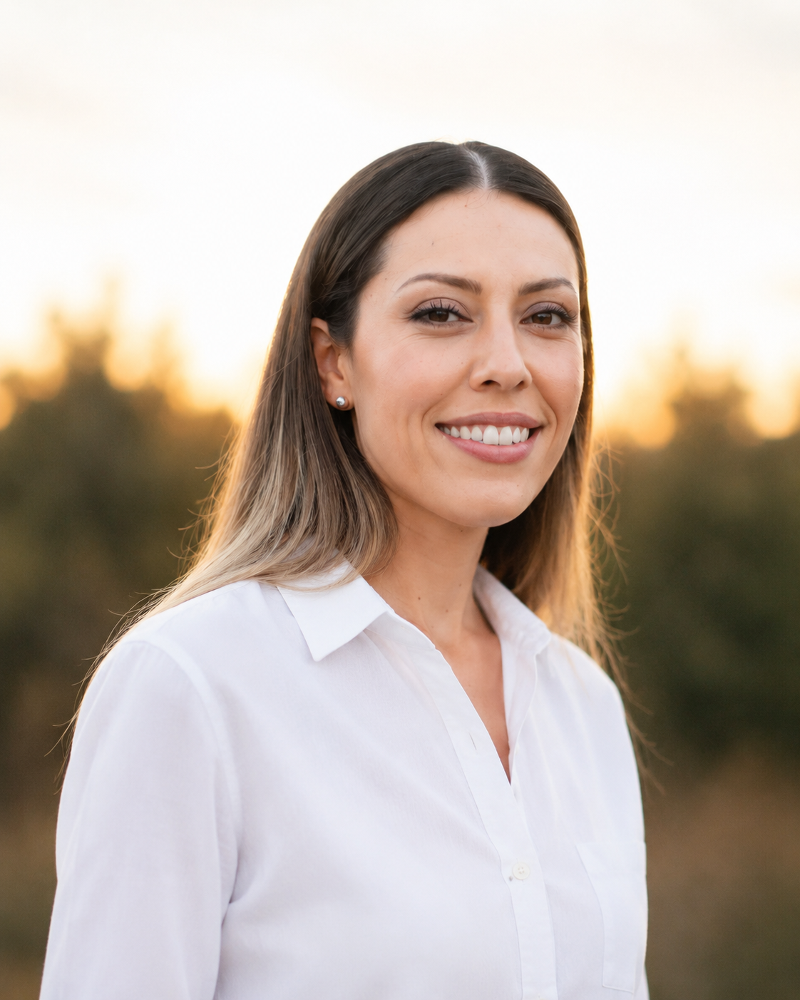 A smiling young woman with long hair holding a glass of red wine outdoors during sunset, wearing a sleeveless white dress with black patterns, tattoos on her arm, and small pearl earrings.