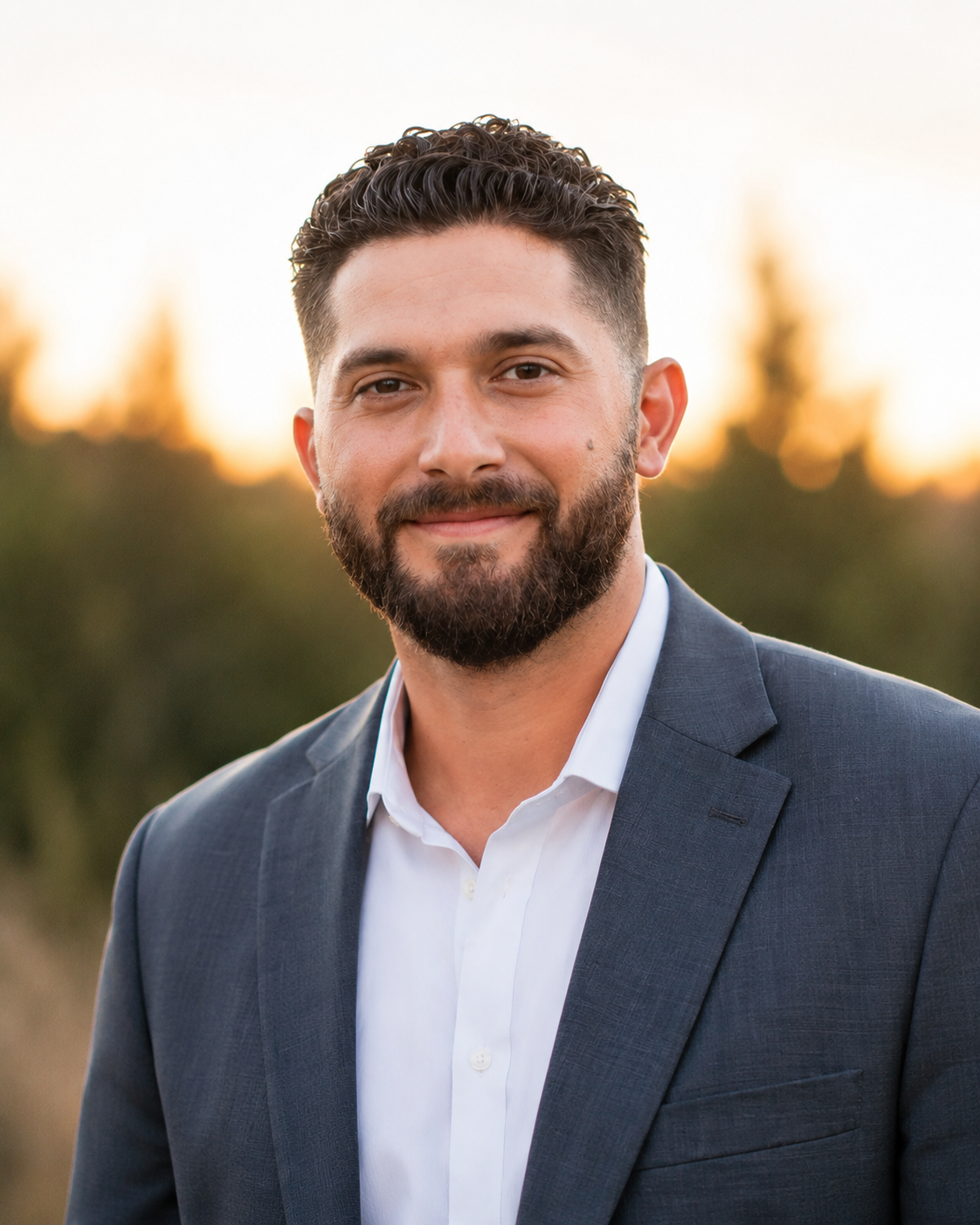 Portrait of a man with a beard and short dark hair, wearing a suit jacket and white dress shirt, outdoors during sunset.