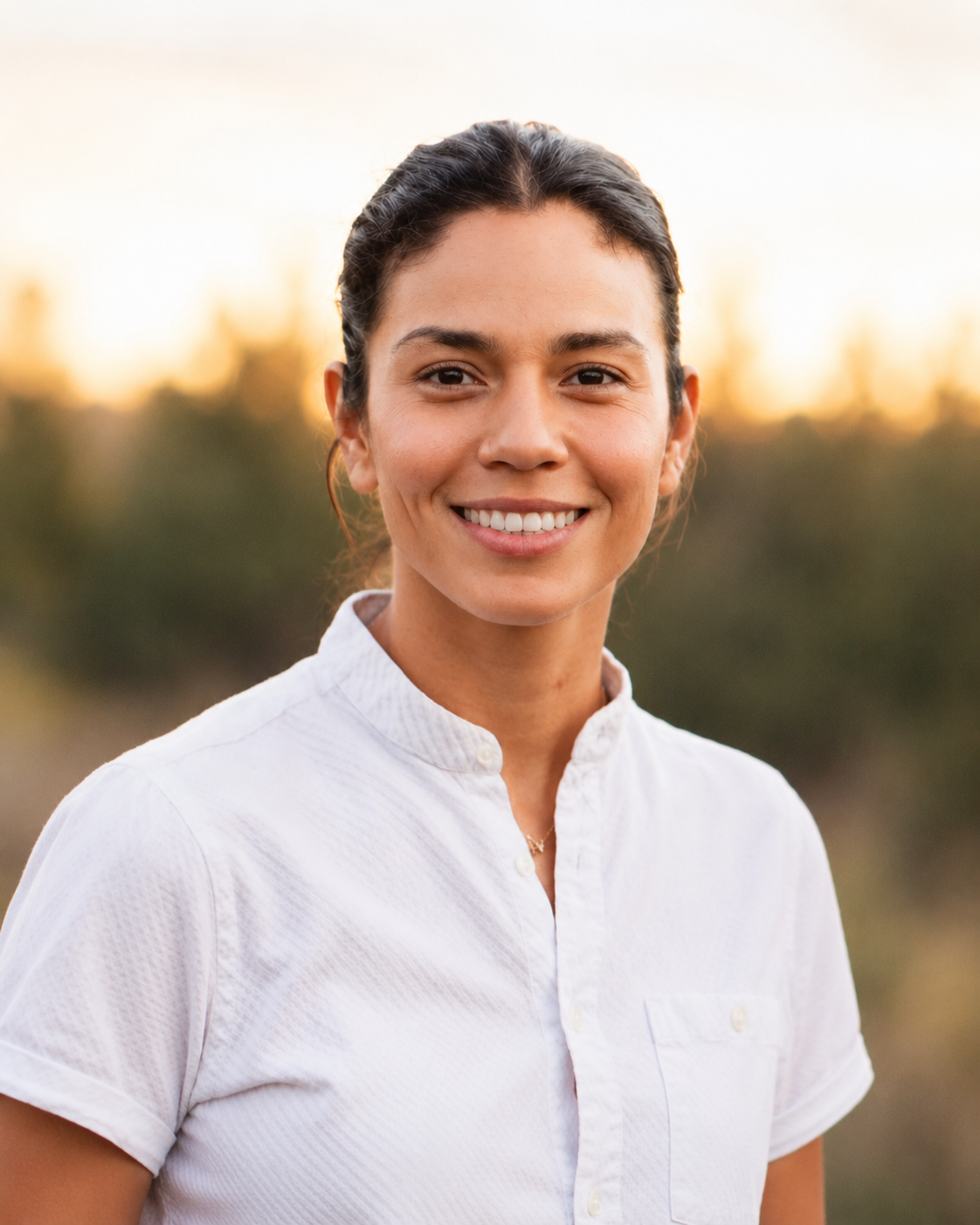 A young woman with dark hair pulled back, smiling, wearing a white shirt, with a mountain landscape in the background during sunset.