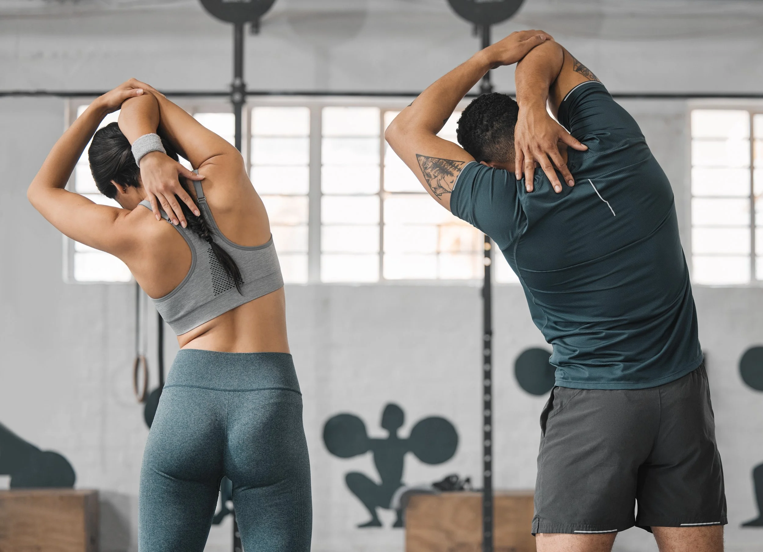Two people in workout clothes stretching their arms overhead in a gym setting.