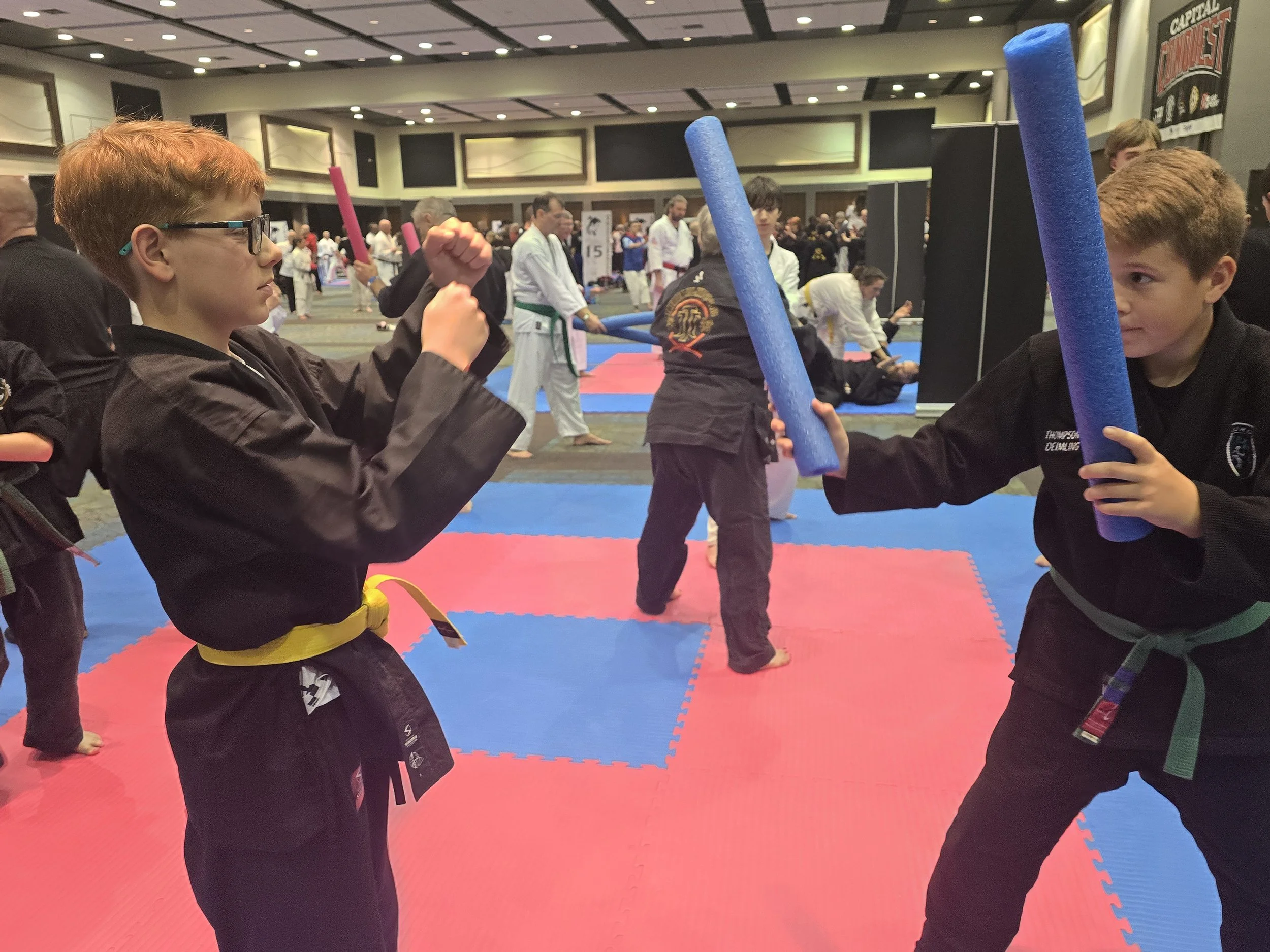Two young boys wearing martial arts uniforms and yellow belts practicing martial arts with foam sticks in a large indoor gym. Other martial artists and spectators are visible in the background.
