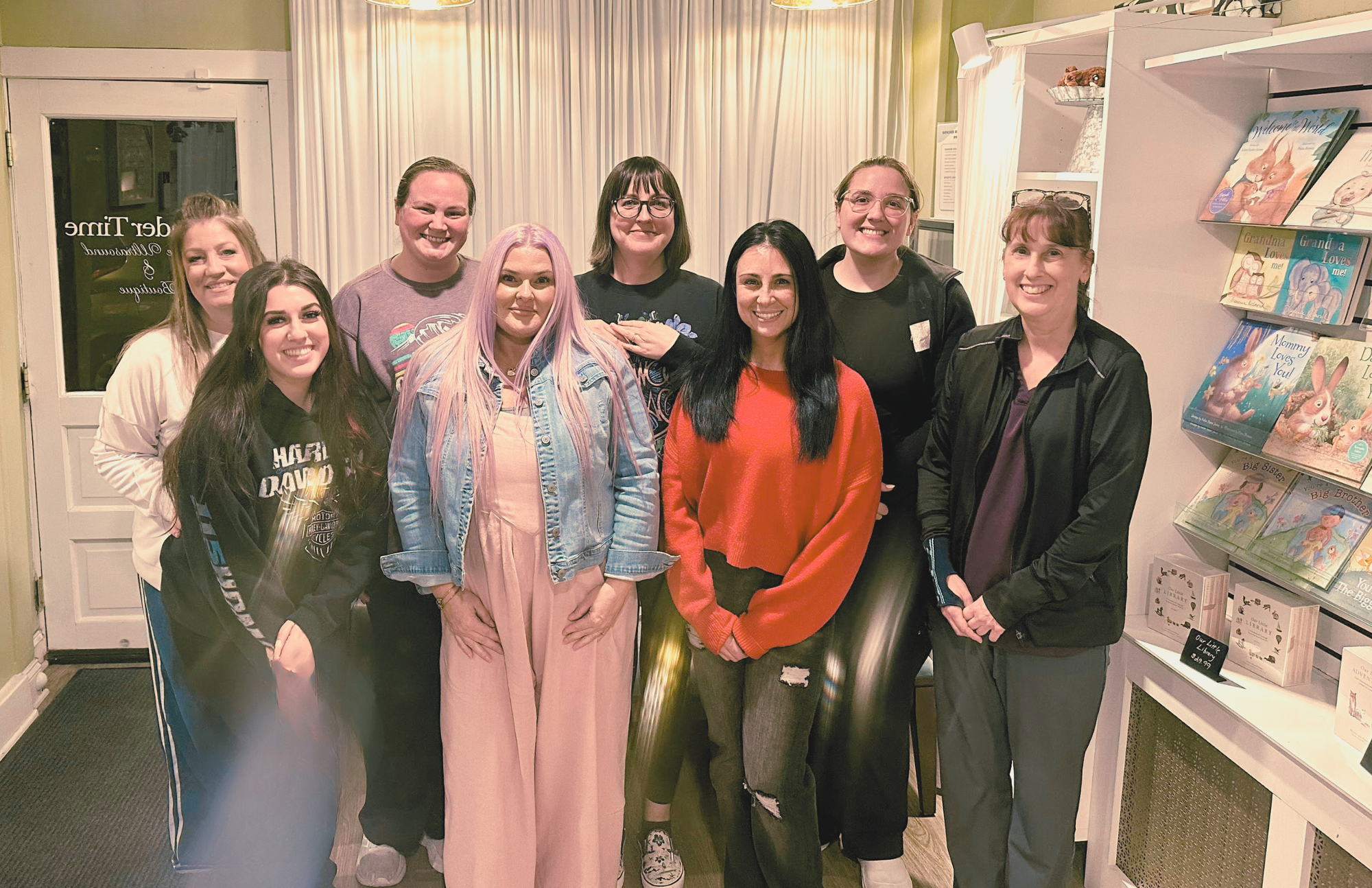 A group of nine women standing inside a bookstore, smiling for a photo. Bookshelves with children's books are visible on the right, and a door with glass is on the left.