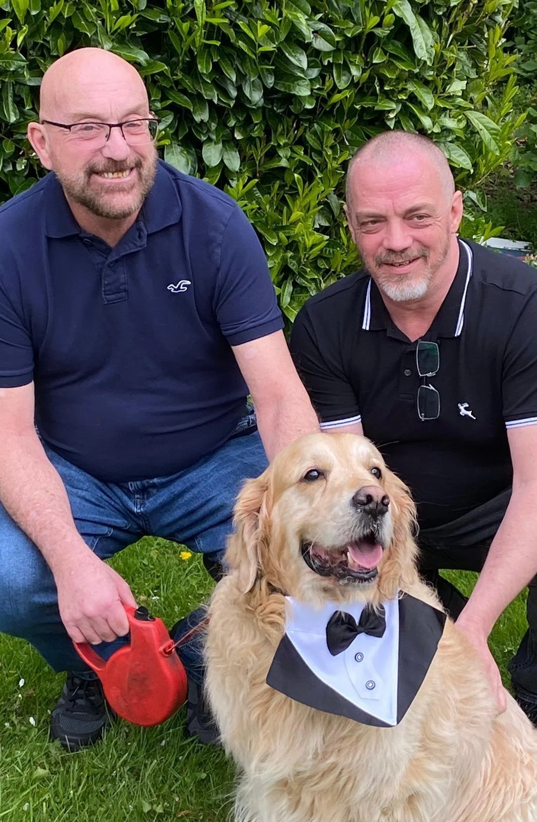 Two smiling men pose outdoors with a golden retriever wearing a tuxedo-themed bandana. One man holds a red leash. Family Recovery.