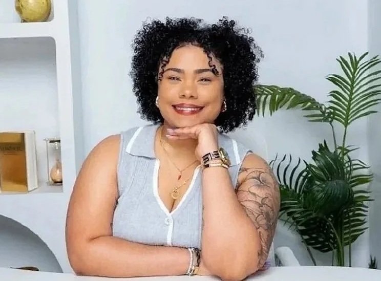 A woman with curly black hair smiling and resting her chin on her hand, sitting at a table with a background of white shelves, books, and green plants.