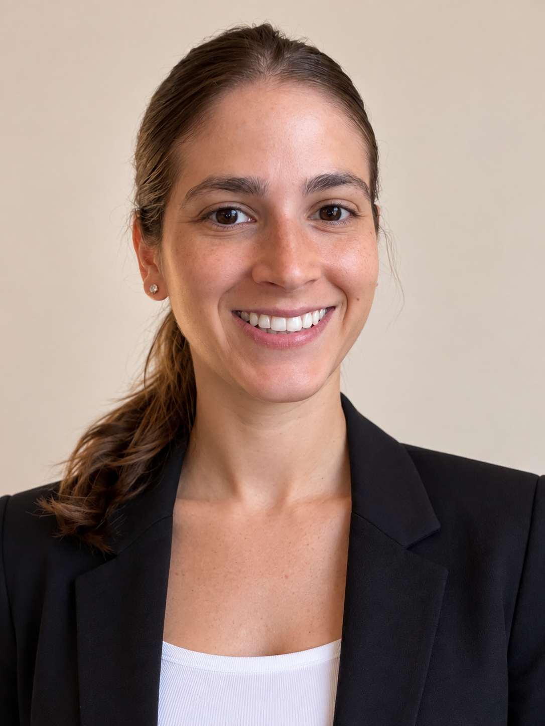 A woman with brown hair tied back, wearing a black blazer and white top, smiling against a plain beige background.