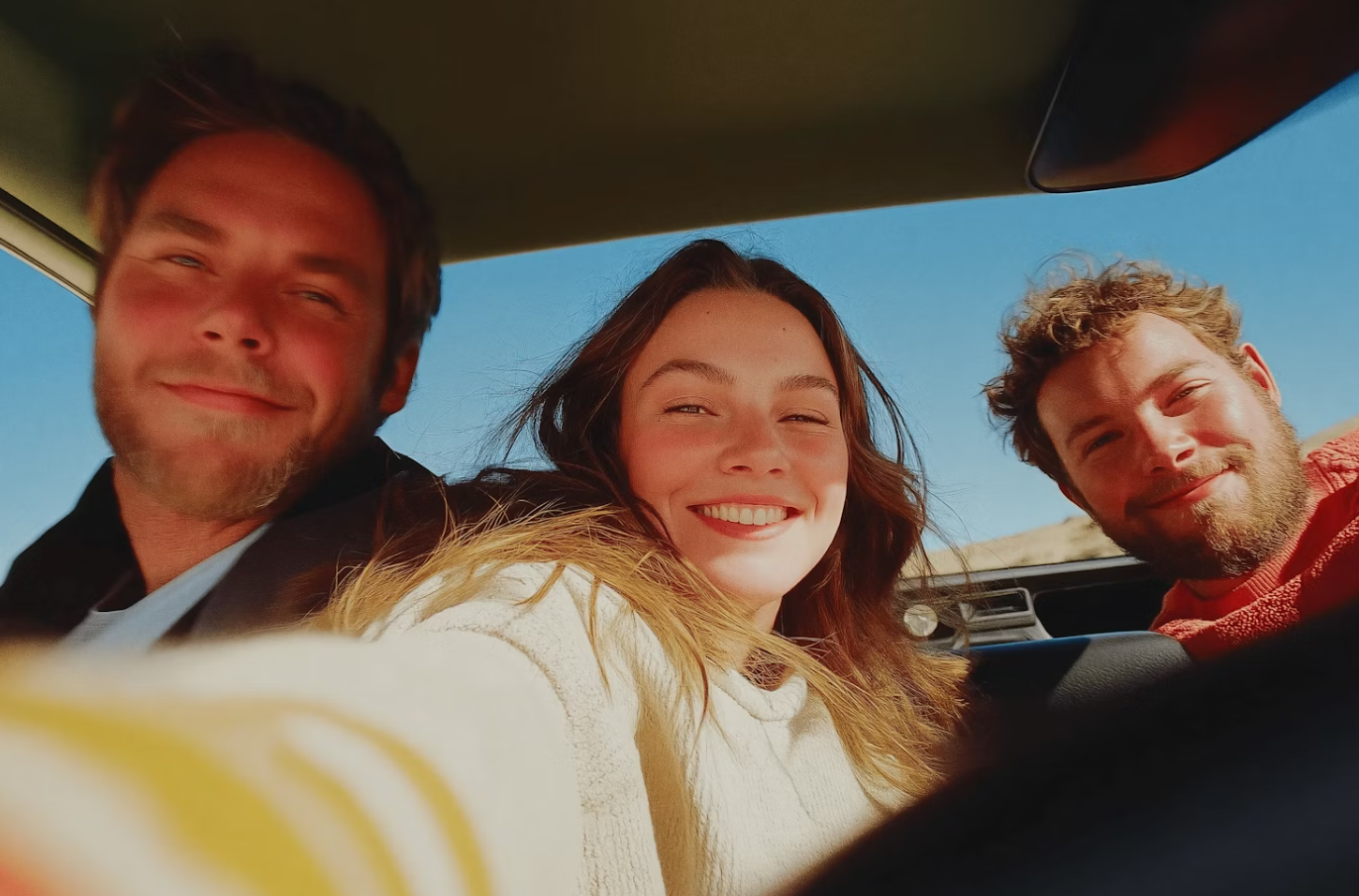 Three friends smiling and taking a selfie inside a car with a clear blue sky outside.