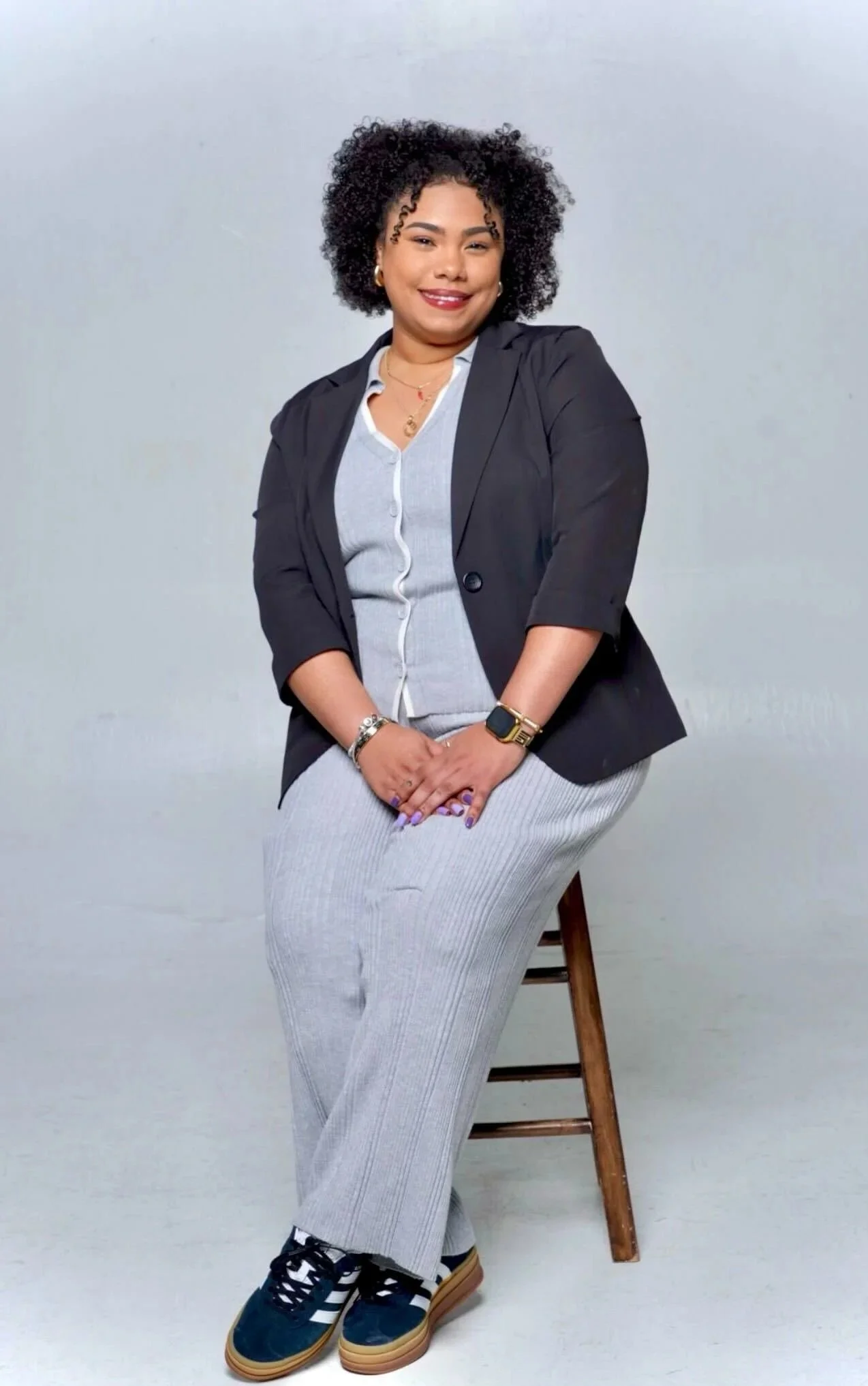 A woman with curly black hair sitting on a stool, dressed in gray pants, a gray button-up shirt, a black blazer, and sneakers, smiling at the camera against a plain background.