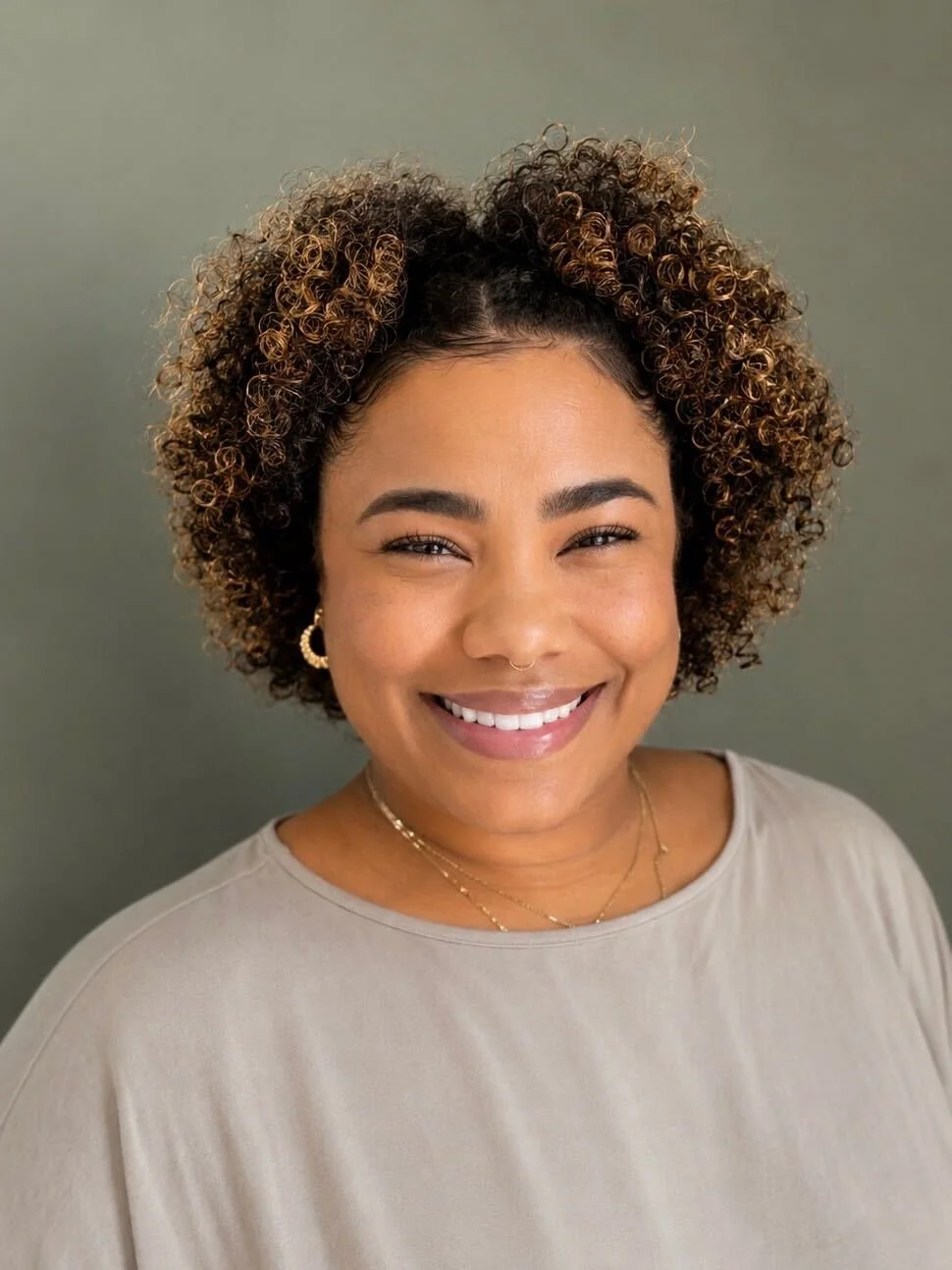 A woman with curly hair smiling, wearing earrings and layered necklaces, against an olive green background.
