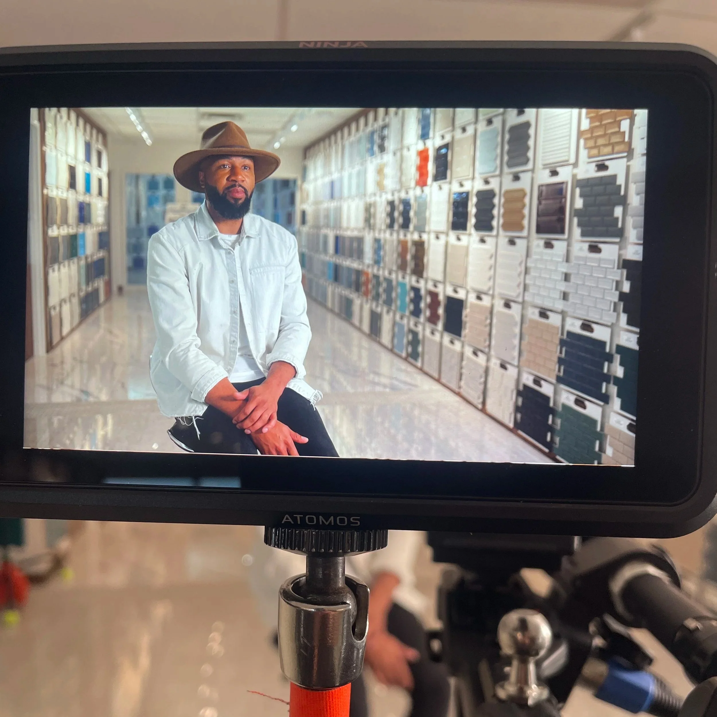 View through a camera display showing a man with a beard wearing a large brown hat and a white shirt, sitting in a showroom with various paint color samples on the wall behind him.