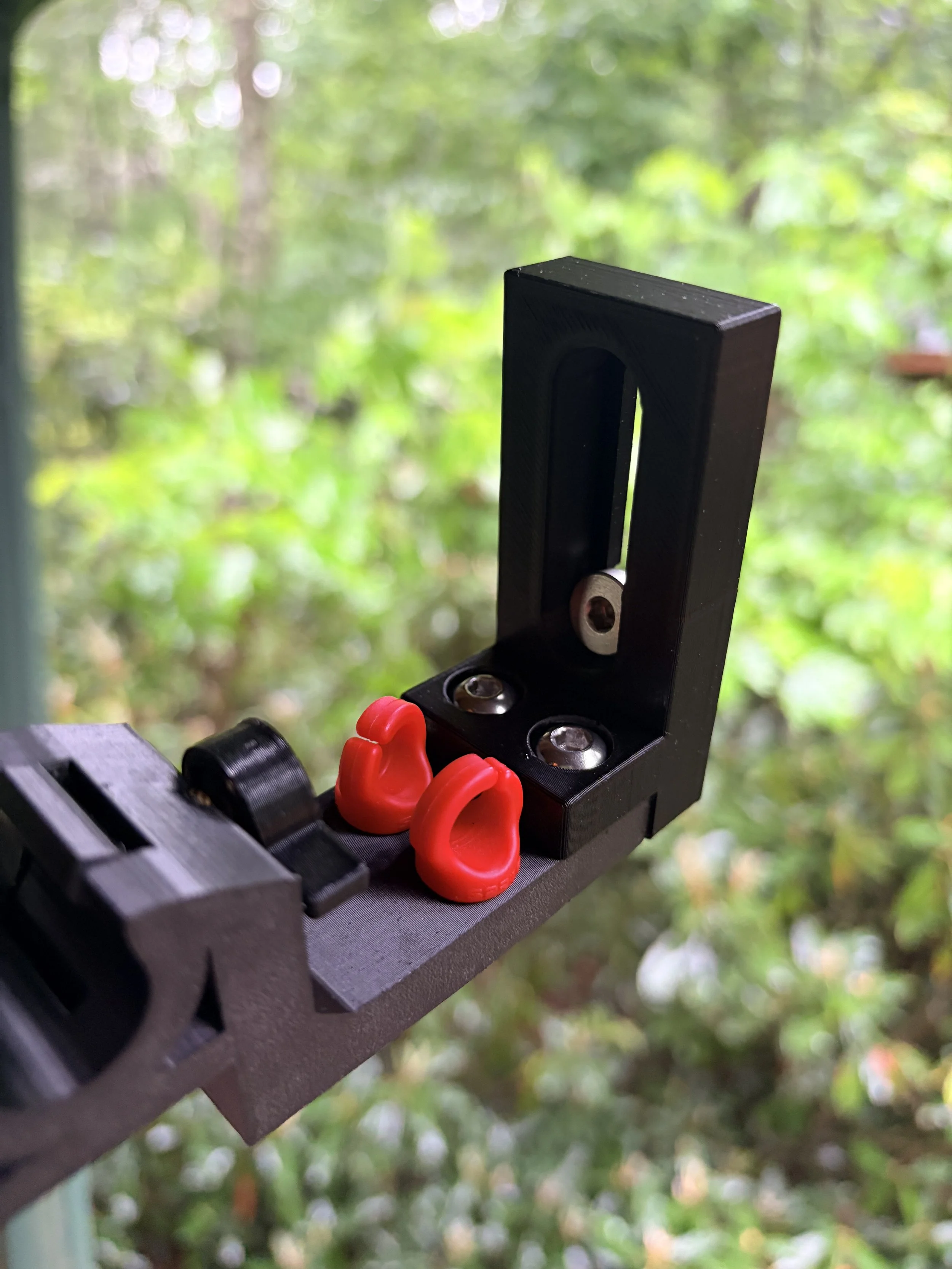 Close-up of a black metal mechanism with red plastic hooks attached, set against a blurred background of green foliage.