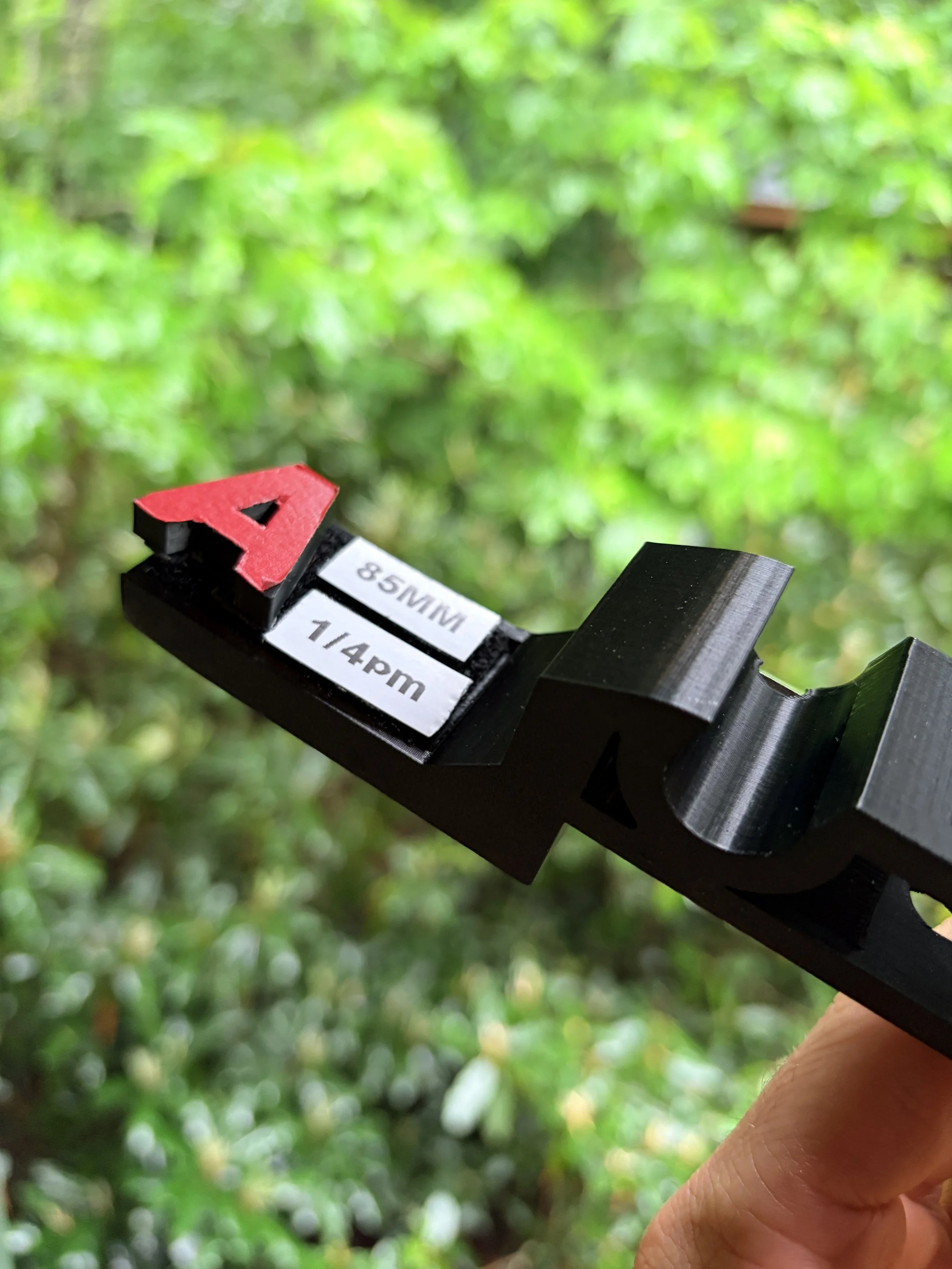 Close-up of a black camera or measurement device with a red arrow-shaped marker and a label indicating 85 millimeters and 1/4 inch, being held by a person outdoors against a green leafy background.