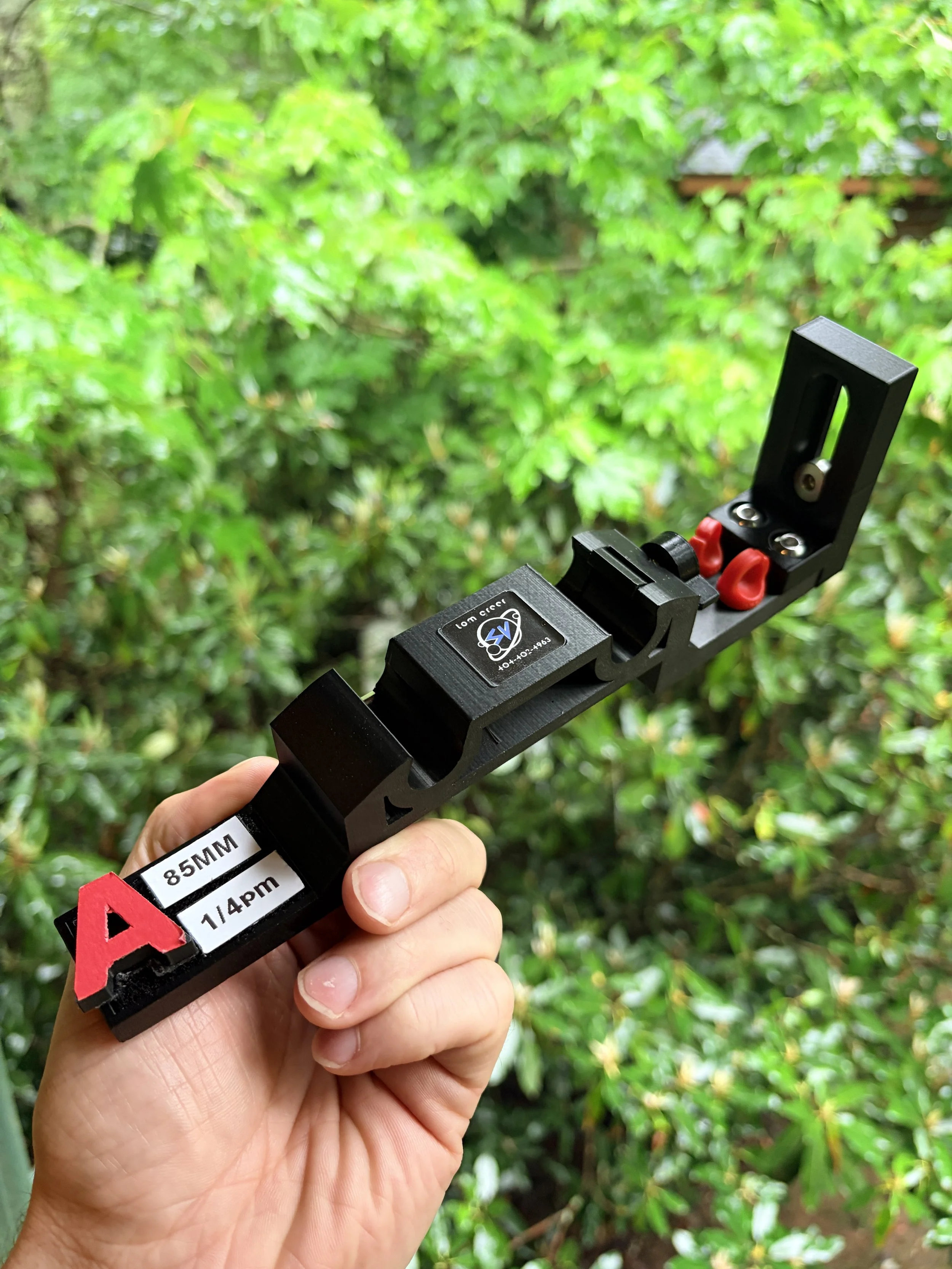 A person holding a black metal rock clamp with a green leafy tree background.