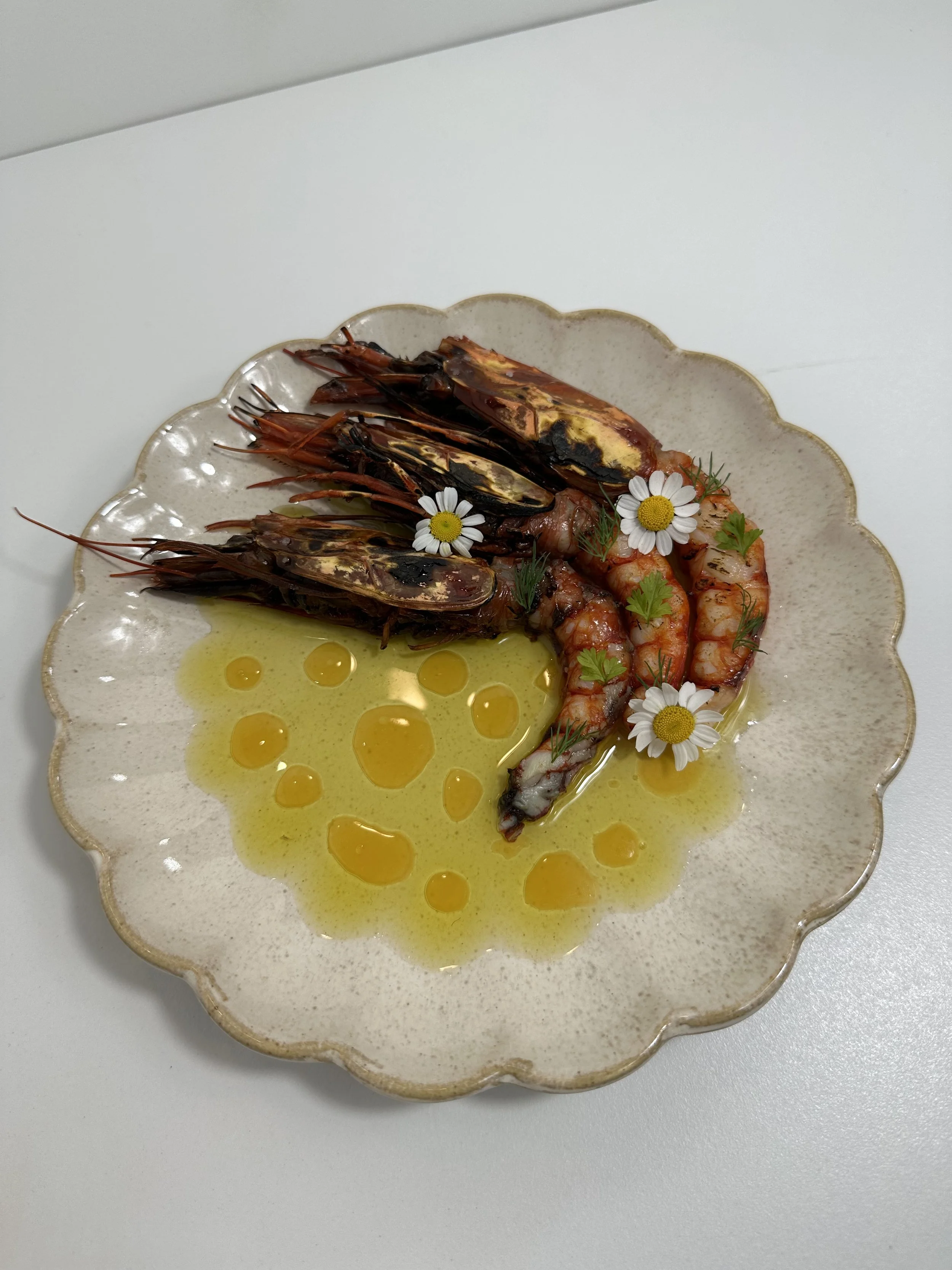 A plate with grilled shrimp, grilled whole prawn, and grilled shellfish garnished with small chamomile flowers and herbs, with olive oil drizzle.