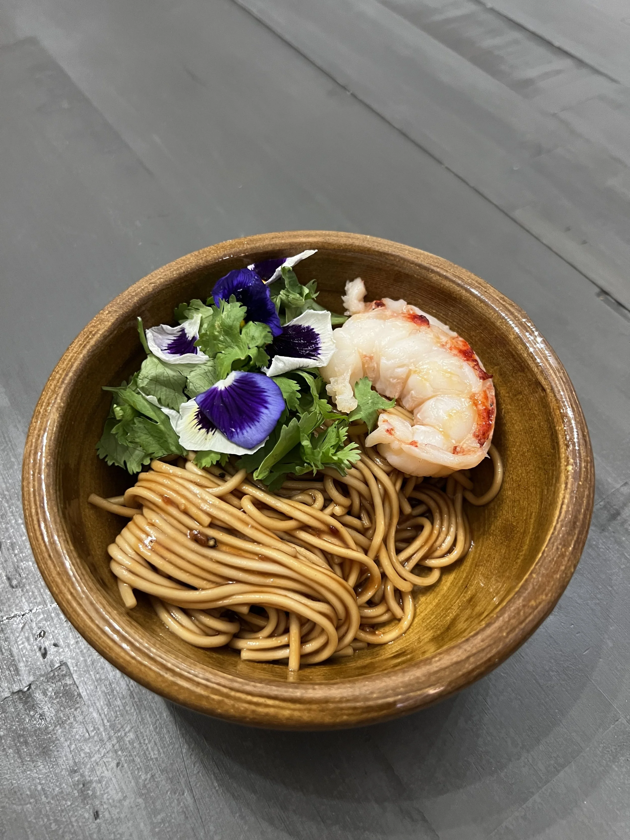 A wooden bowl with noodles, a cooked shrimp, fresh greens, and edible flowers on top, placed on a gray table.