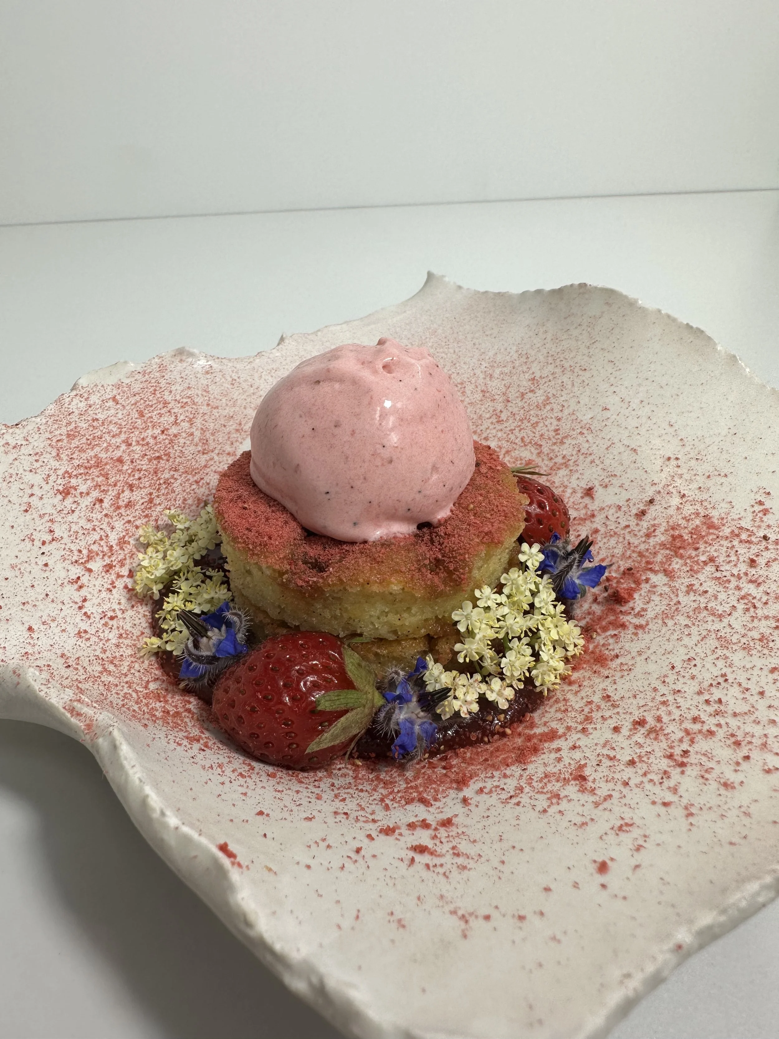 Dessert plate with a slice of cake topped with pink ice cream, strawberries, edible flowers, and pink powder on a decorative white ceramic plate.
