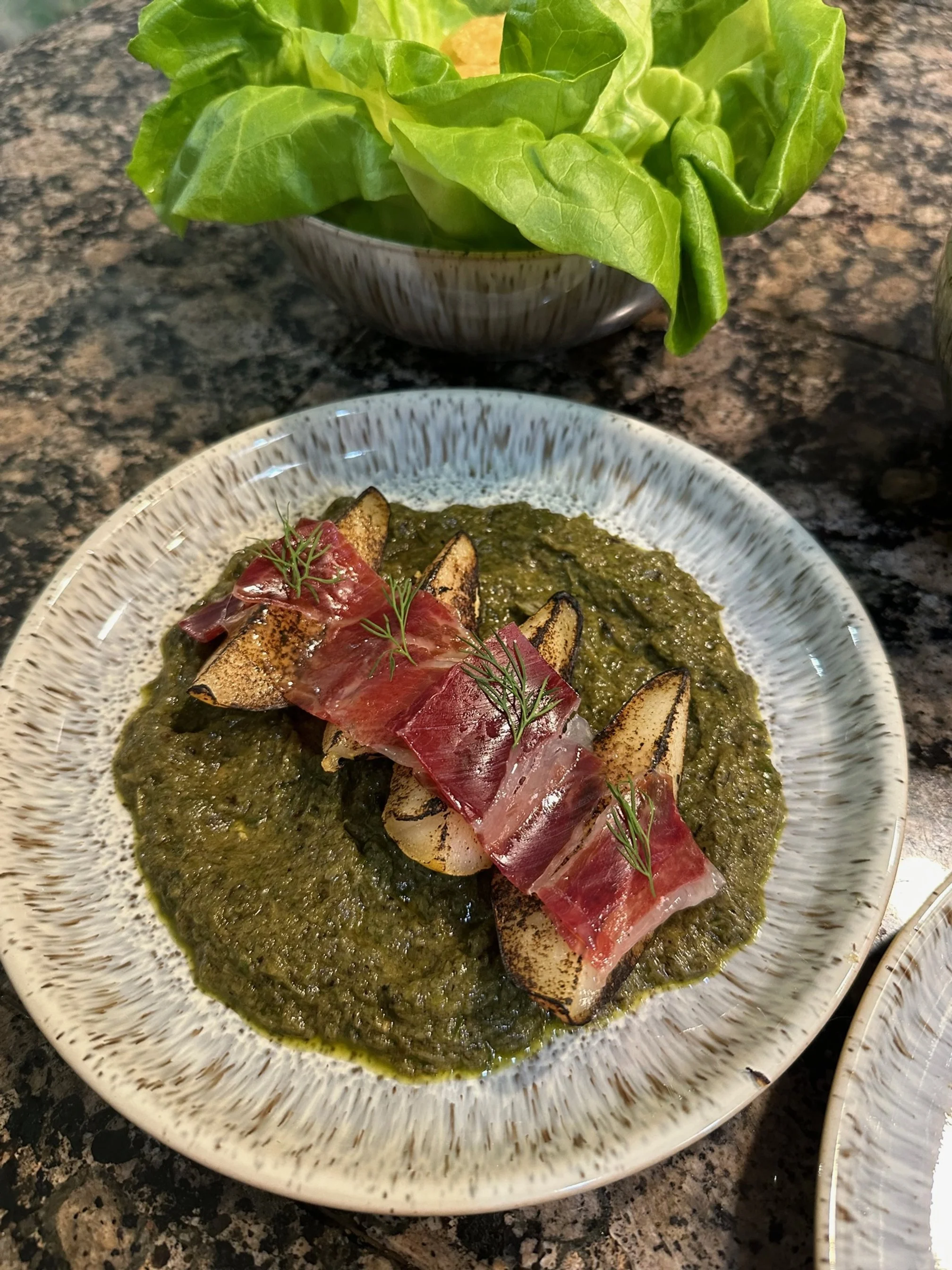 Grilled eggplant slices topped with raw prosciutto, garnished with fresh herbs, served on a bed of green sauce with a side of lettuce salad.