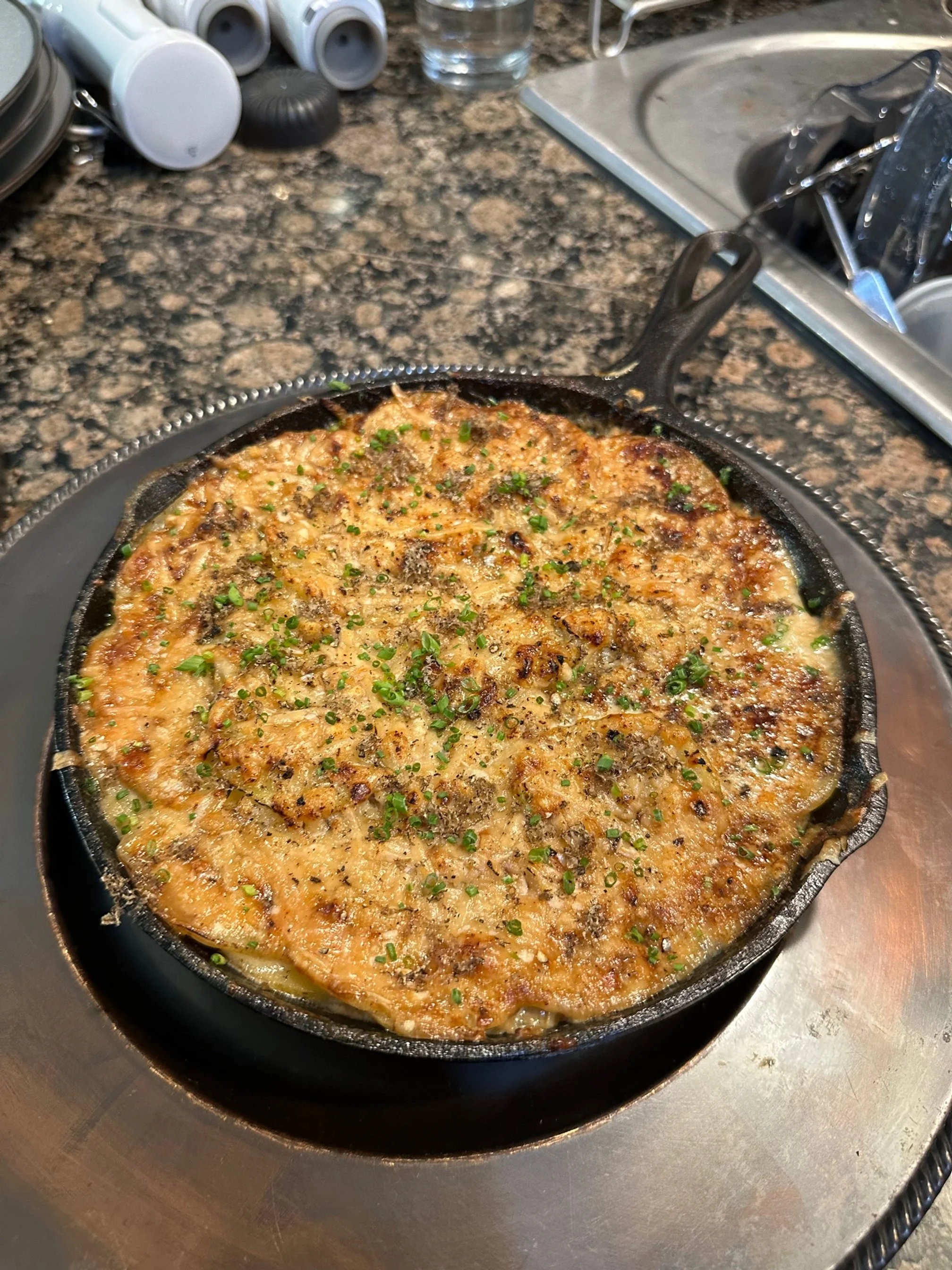 A skillet of baked macaroni and cheese topped with cheese, herbs, and seasoning, set on a metal tray on a kitchen countertop.