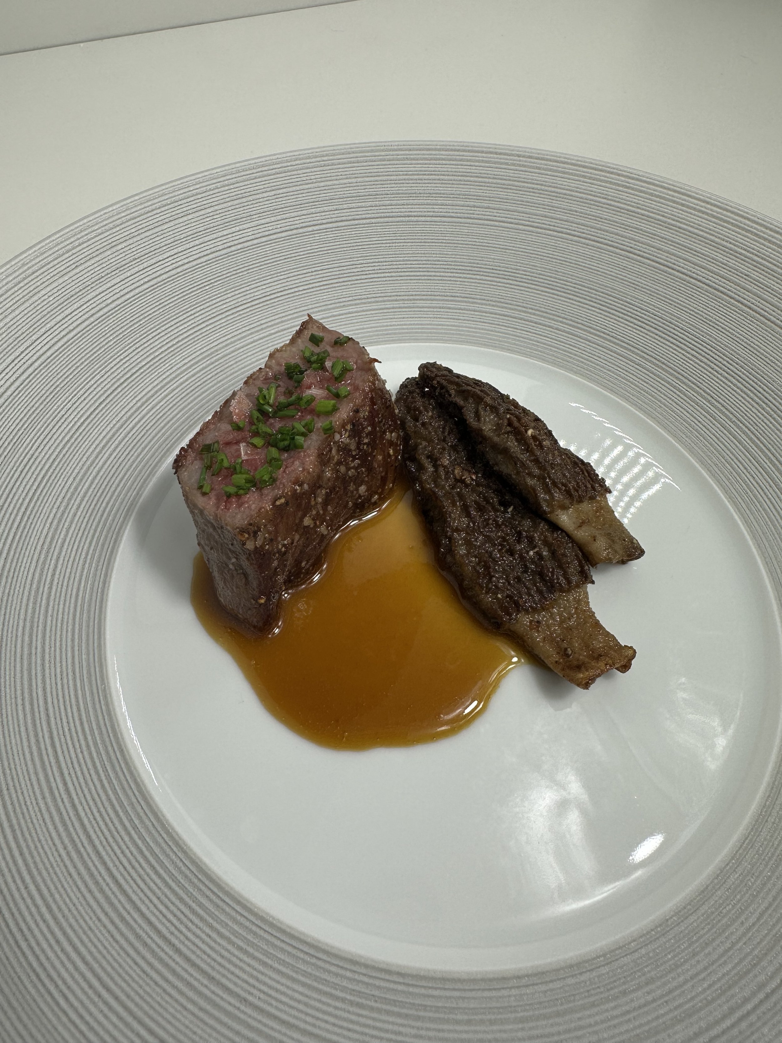 High-end plated dish featuring a rectangular cut of seared steak with green herbs on top, and two dark, crispy-looking ribs, served with a rich brown sauce on a white plate with a textured silver rim.