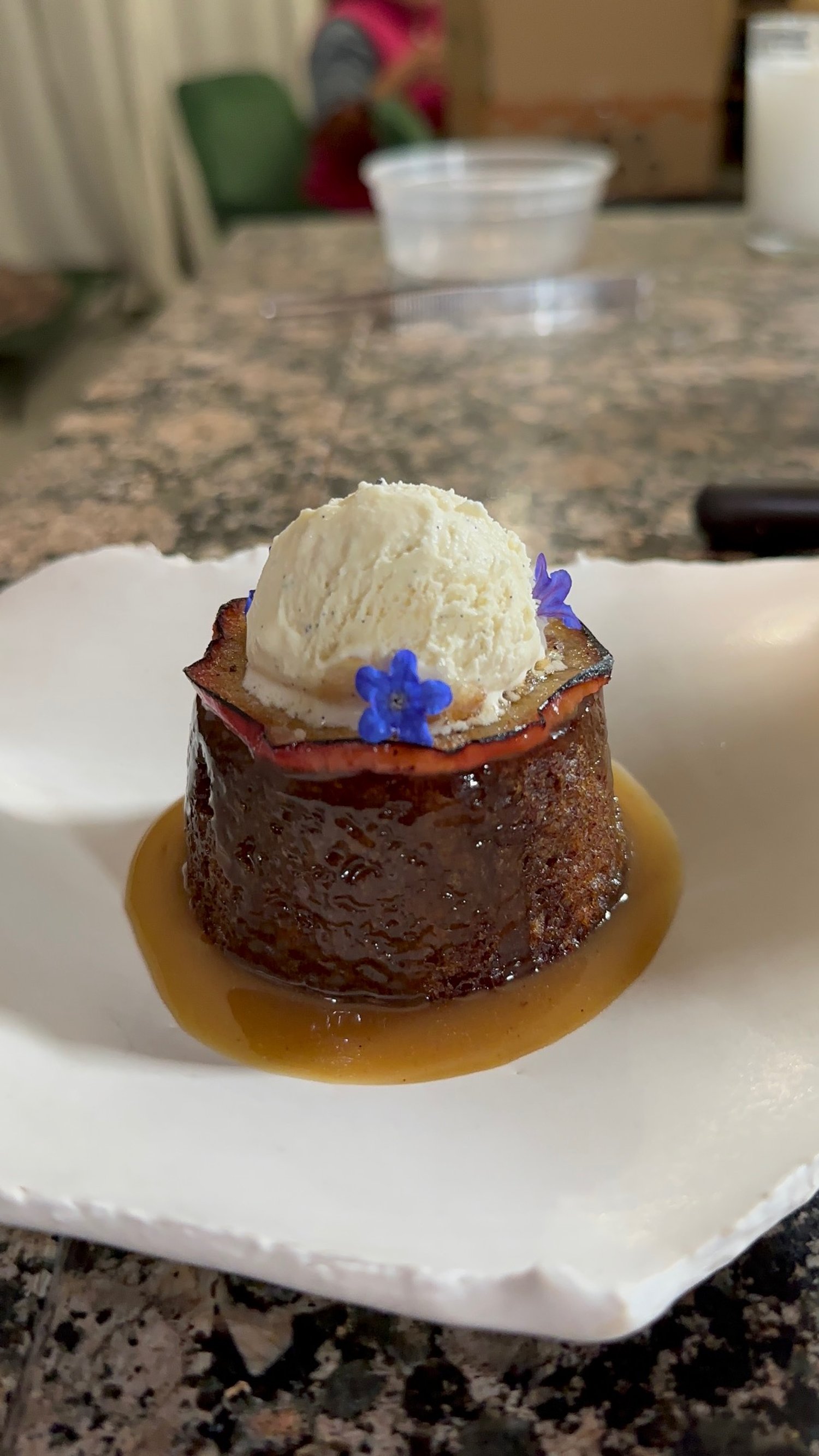 A dessert with a caramelized flan topped with a small scoop of vanilla ice cream, a crispy bacon strip, and blue edible flowers on a white plate.