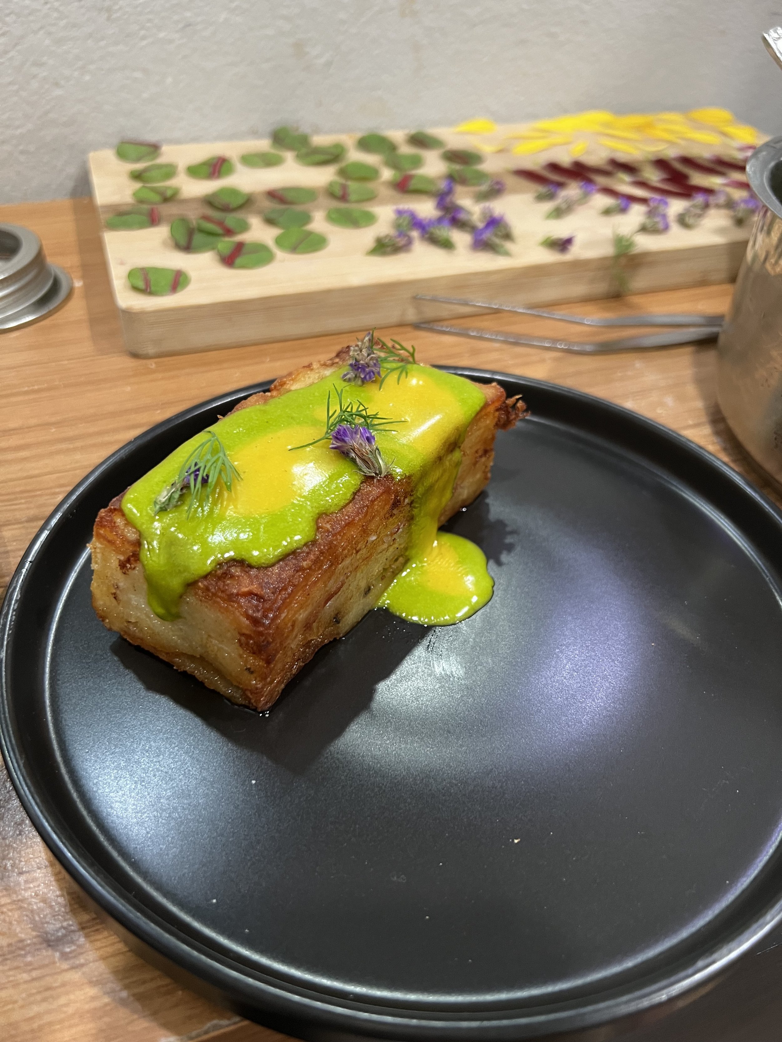 A piece of dessert with a golden brown crust, topped with yellow and green sauces and garnished with small purple flowers and green herbs, served on a black plate.