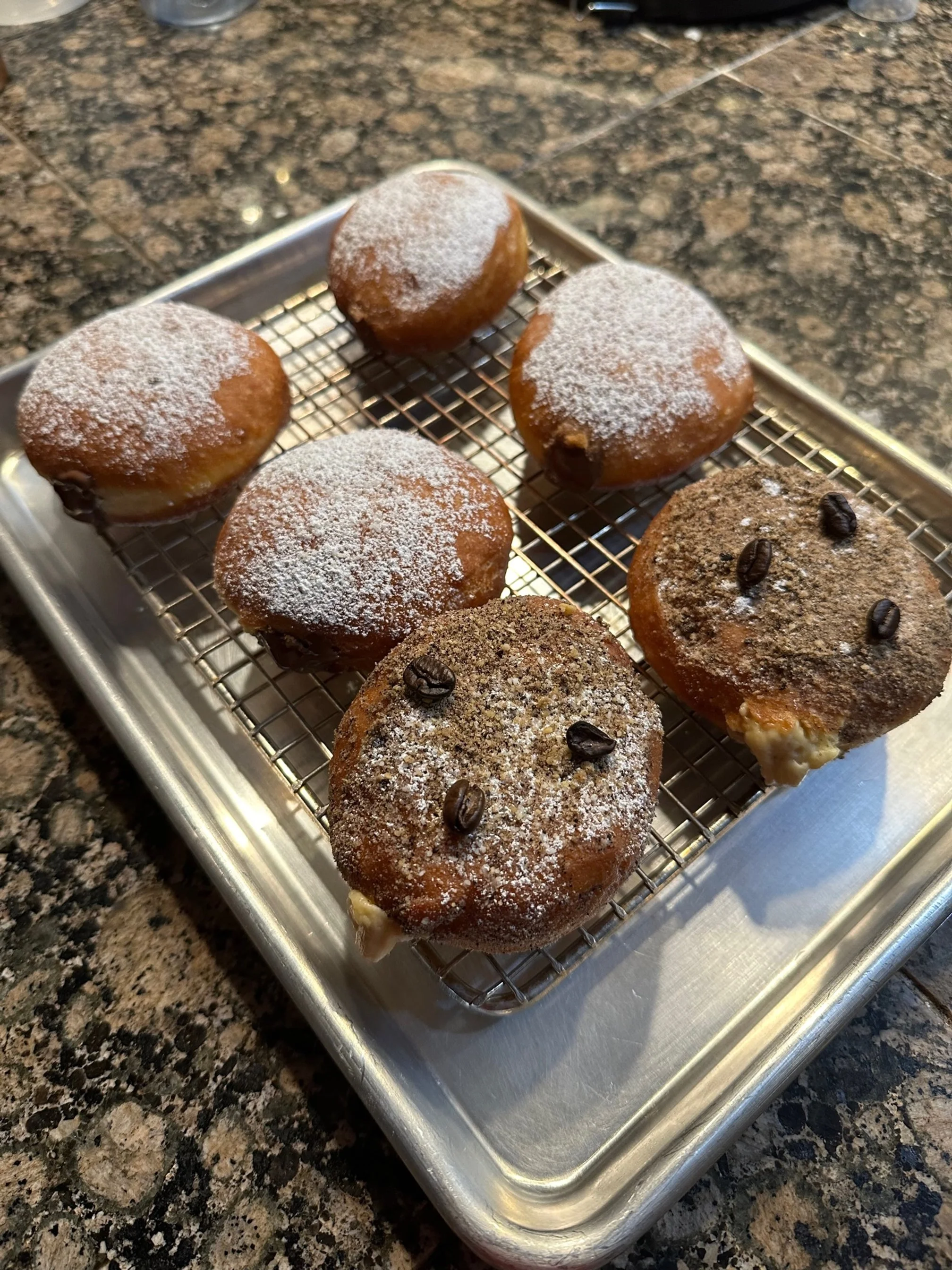 Six donuts on a wire rack, dusted with powdered sugar and topped with coffee beans, on a metal tray.