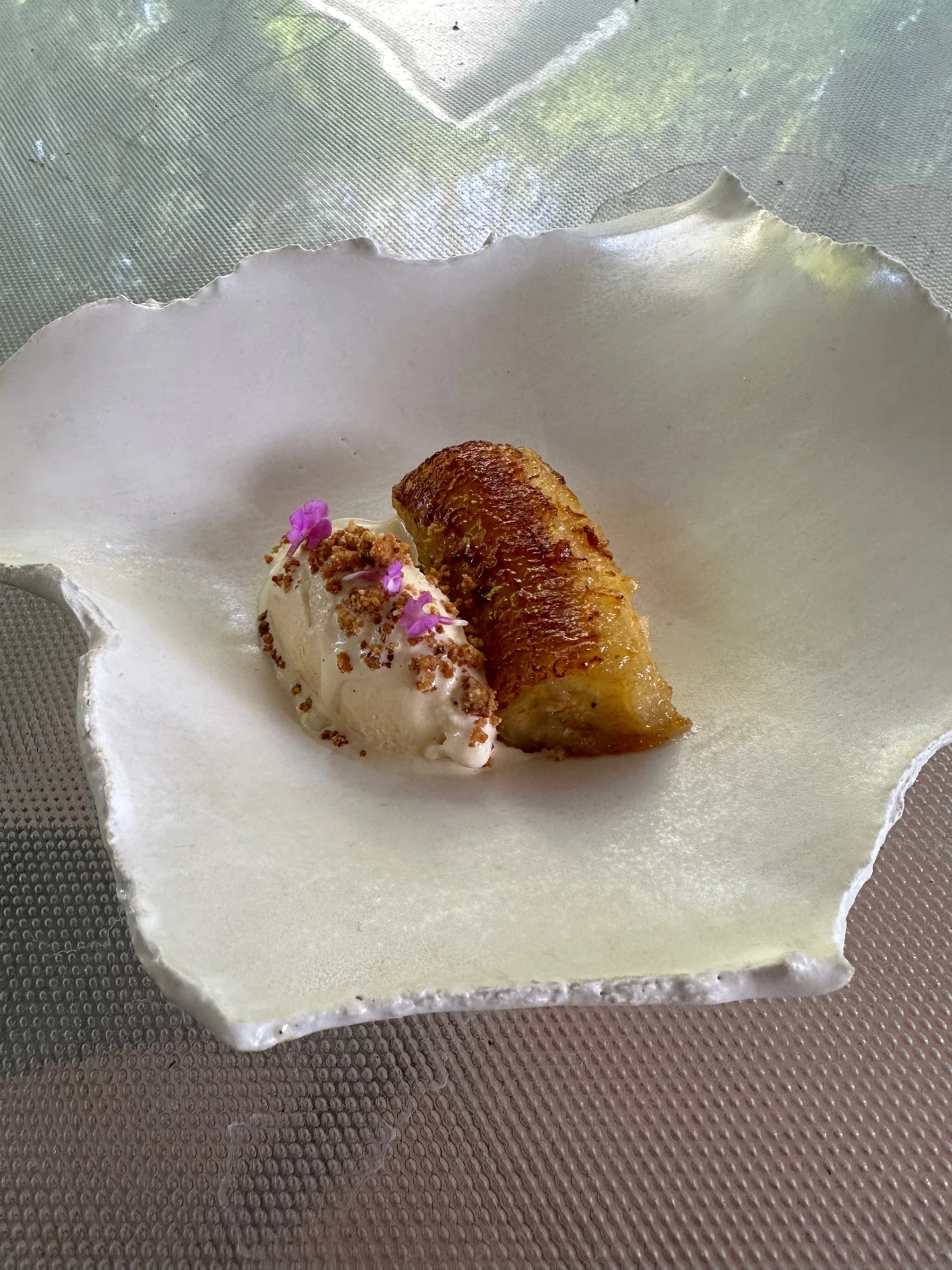 A plated dessert featuring a caramelized banana, a scoop of vanilla ice cream garnished with pink edible flowers and crumbled toppings, served on a white, textured, wavy ceramic plate.