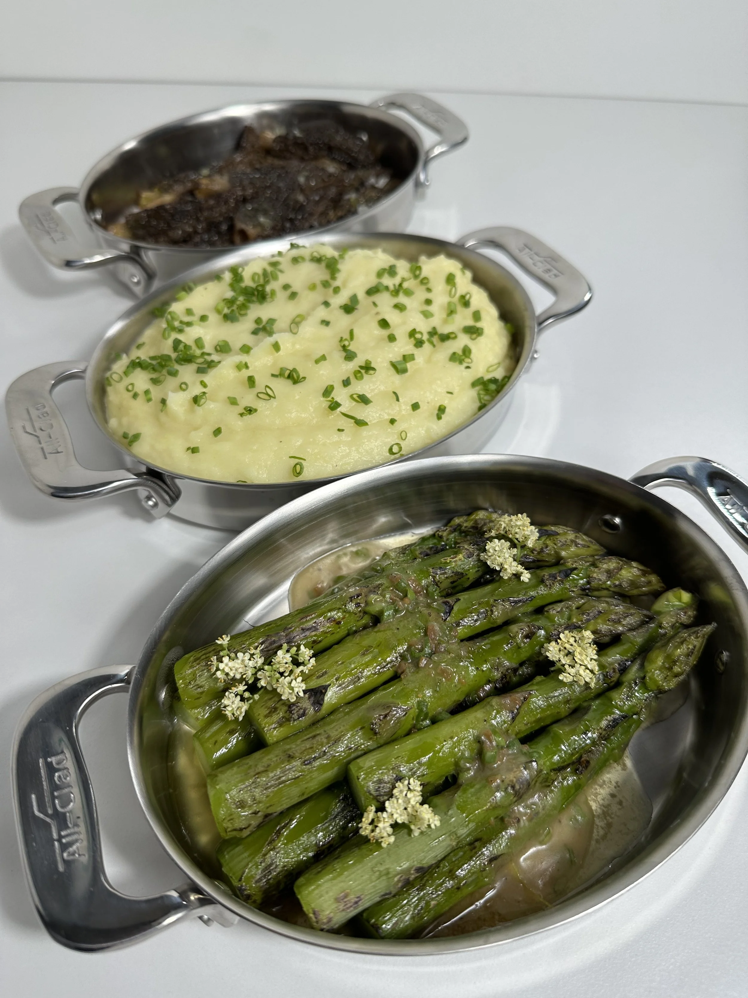 Three small stainless steel dishes containing cooked food, including grilled steak, mashed potatoes with chives, and roasted asparagus with edible flowers.