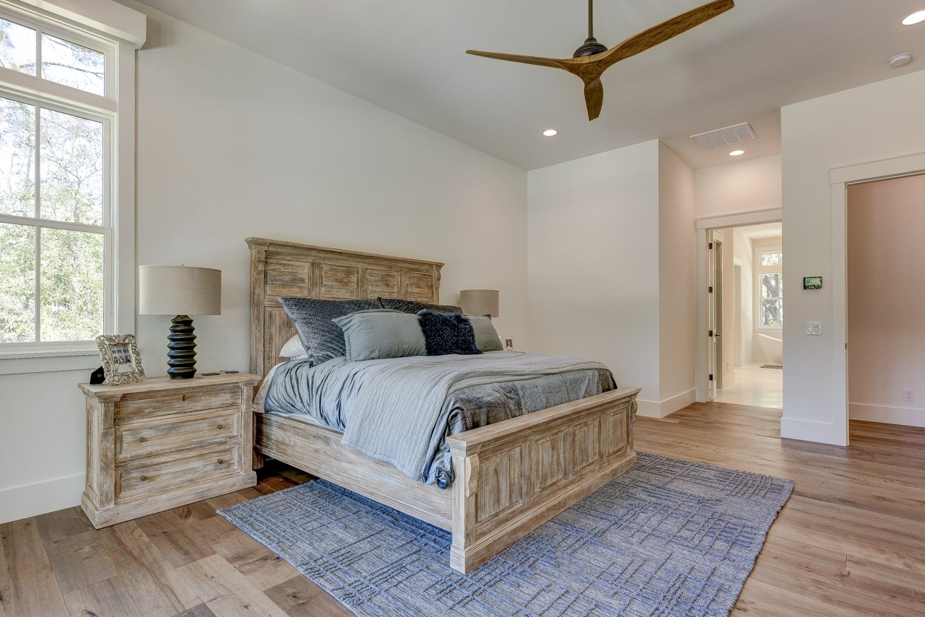 Modern bedroom with wooden bed frame, blue bedding, matching nightstand, lamps, ceiling fan, large window, and hardwood floor.