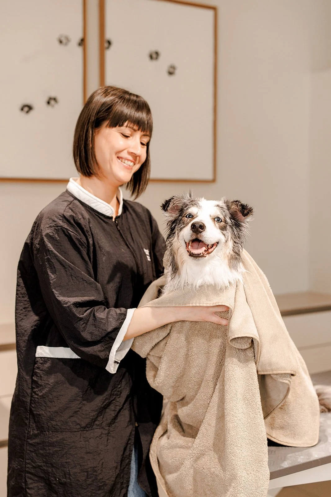 Bark & Bone Dog Grooming  woman with short dark hair and a black jacket smiling as she holds a happy dog wrapped in a towel.
