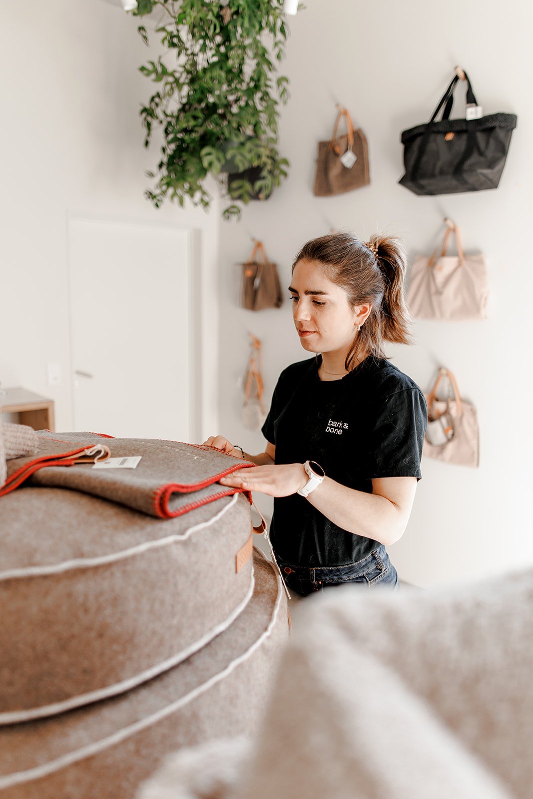 Bark & Bone Dog Daycare Dog Shop A woman with brown hair in a ponytail, wearing a black t-shirt and white watch, is inspecting a brown pet bed with red trim in a retail store, with various handbags hanging on the wall behind her.