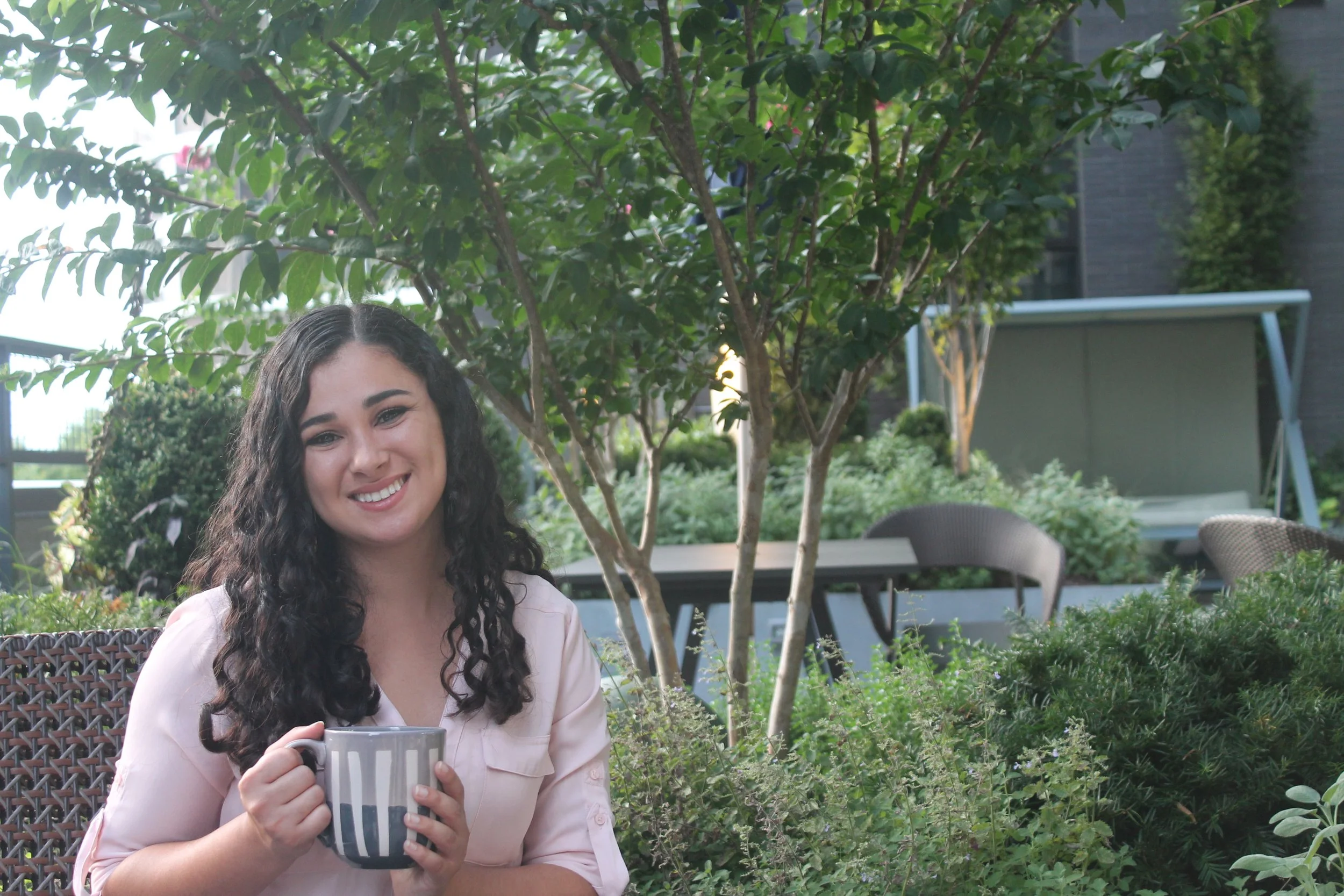 A woman with curly dark hair smiling outdoors while holding a striped mug, surrounded by green plants and garden furniture.