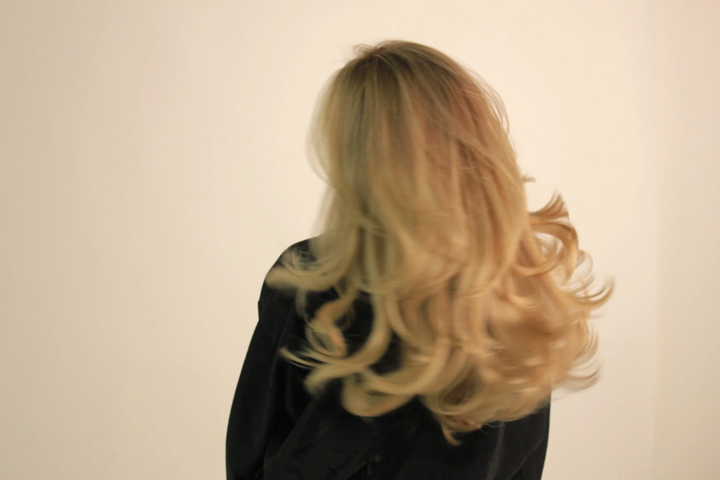 A woman with long, wavy blonde hair wearing a black top standing against a plain light-colored background.