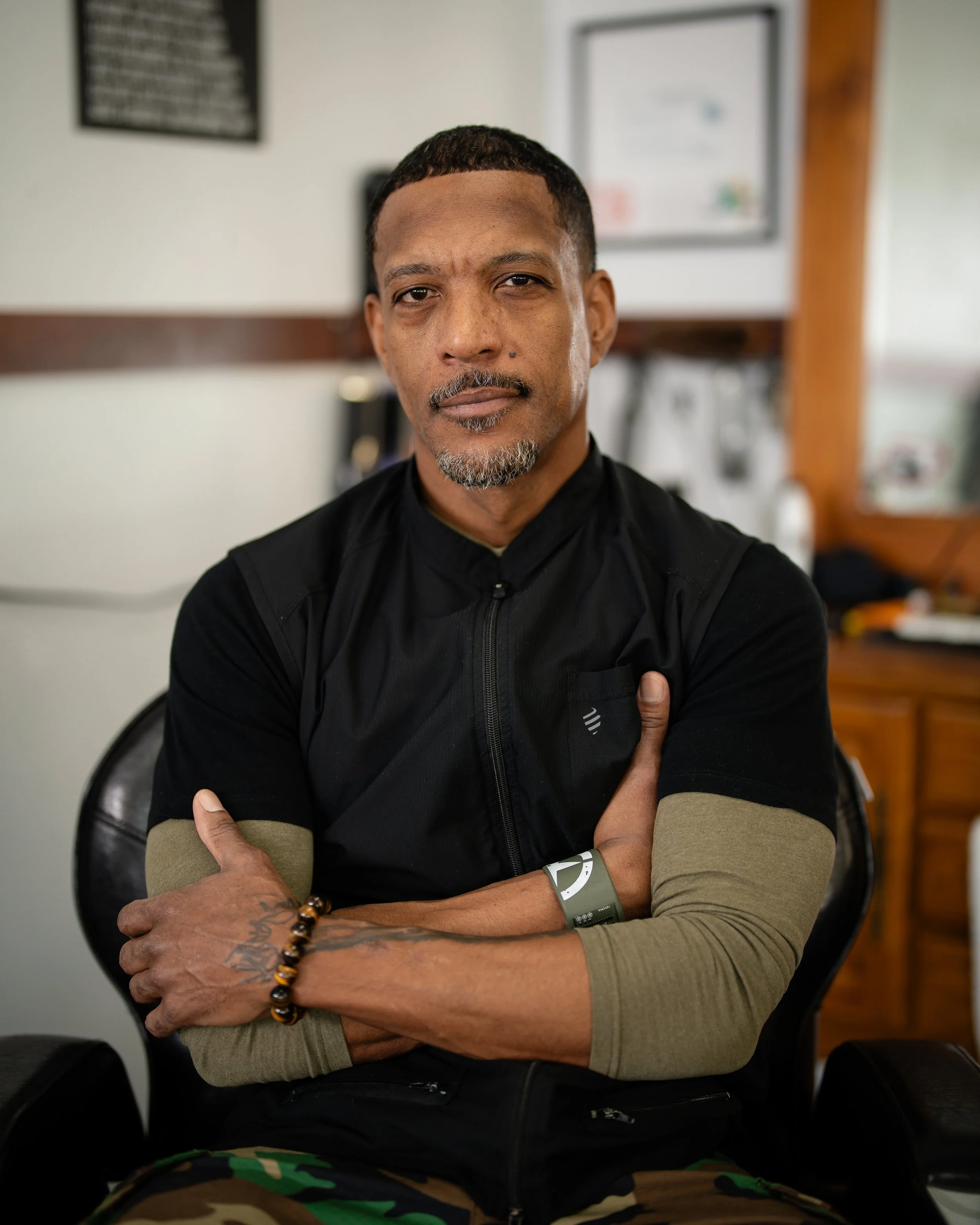 A man with a goatee and tattooed arm holds hairdressing tools in front of a mirror in a modern hair salon. He wears a black cap and vest, with salon equipment and another person reflected in the mirror.