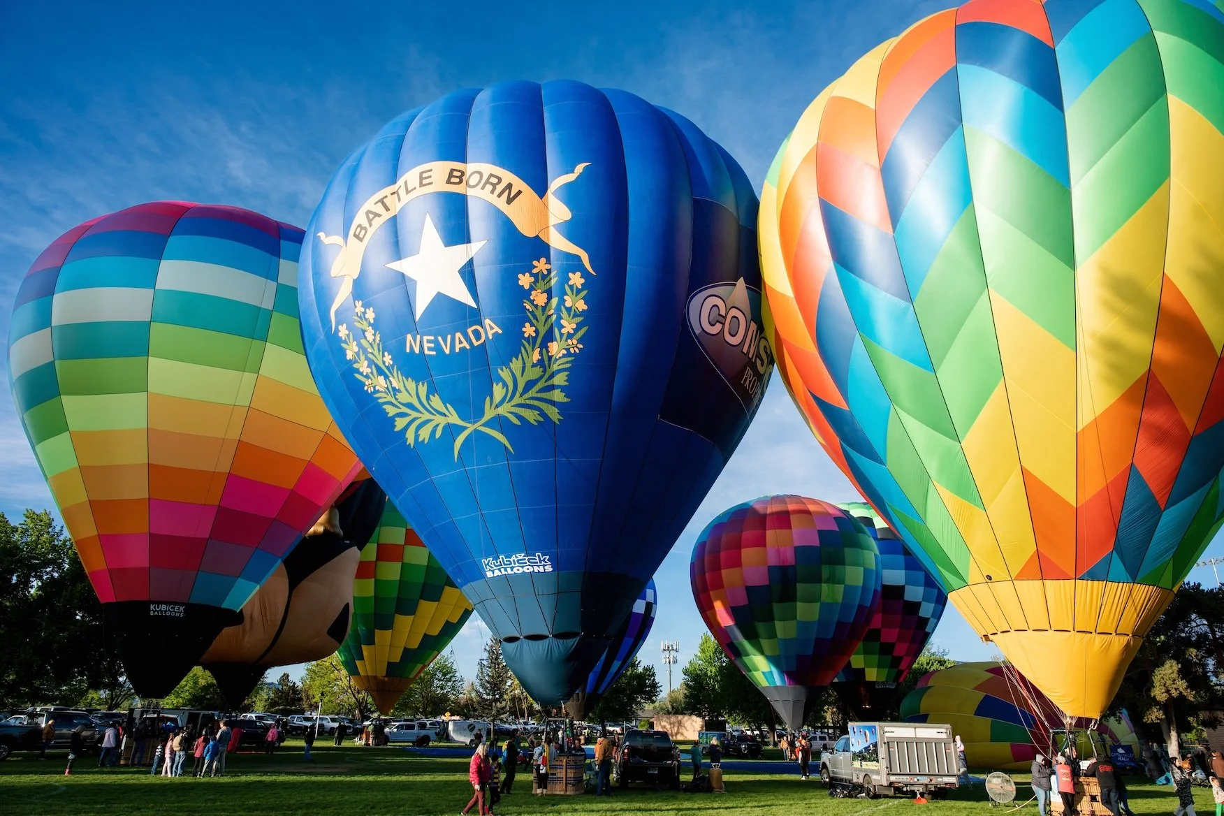 Multiple hot air balloons of various colorful patterns inflated and ready for flight, with people gathered on the ground.