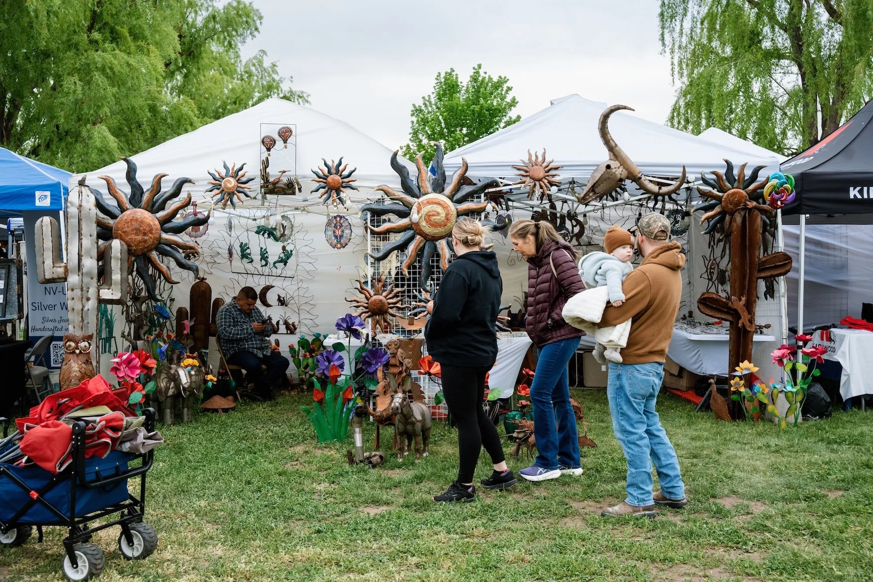 People browsing art and craft items at an outdoor market stall with metal sun and animal sculptures, colorful metal flowers, and onlookers, including a man holding a child, in front of tents and greenery.