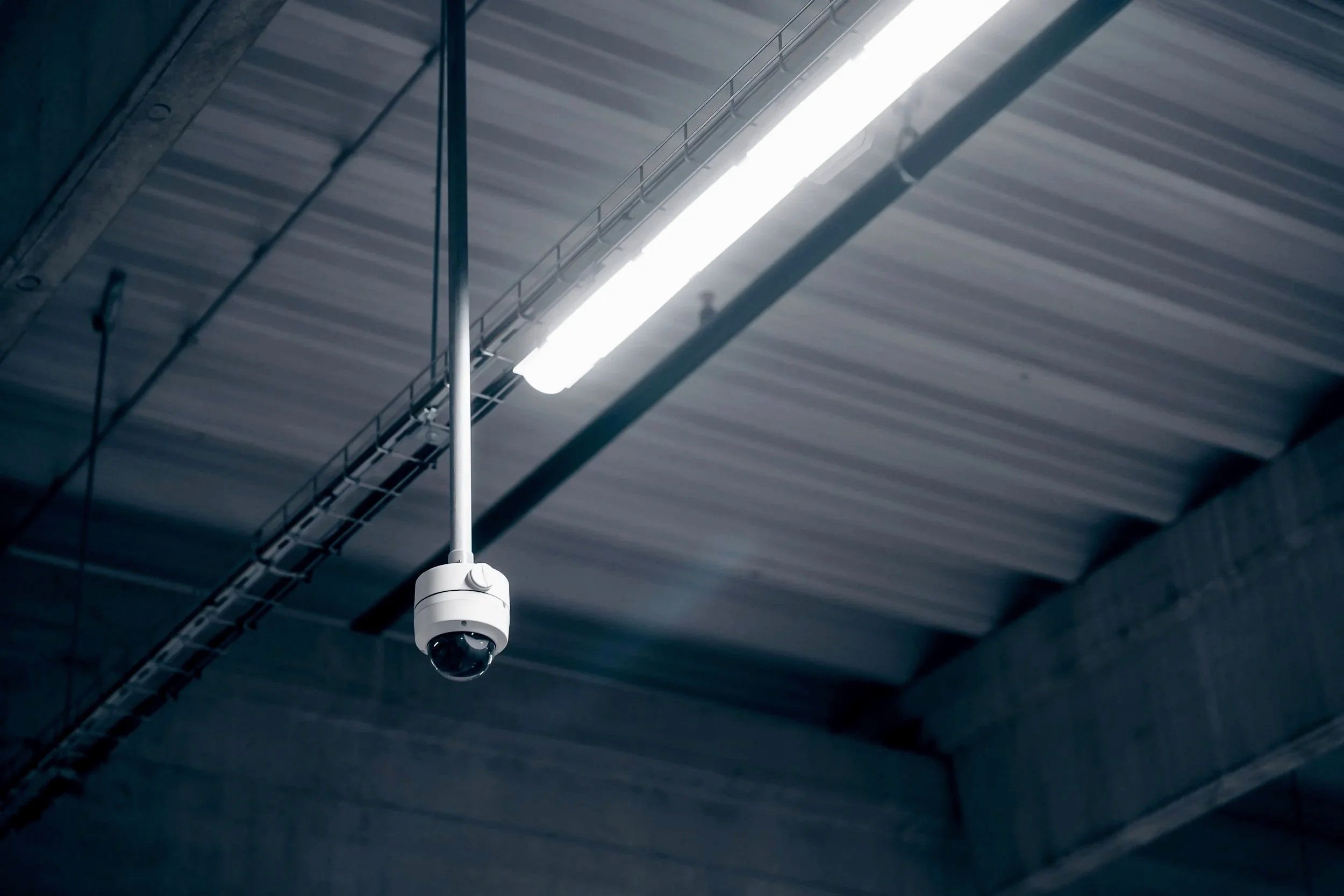 Security camera hanging from the ceiling of an industrial building with exposed metal roof panels and fluorescent lighting.