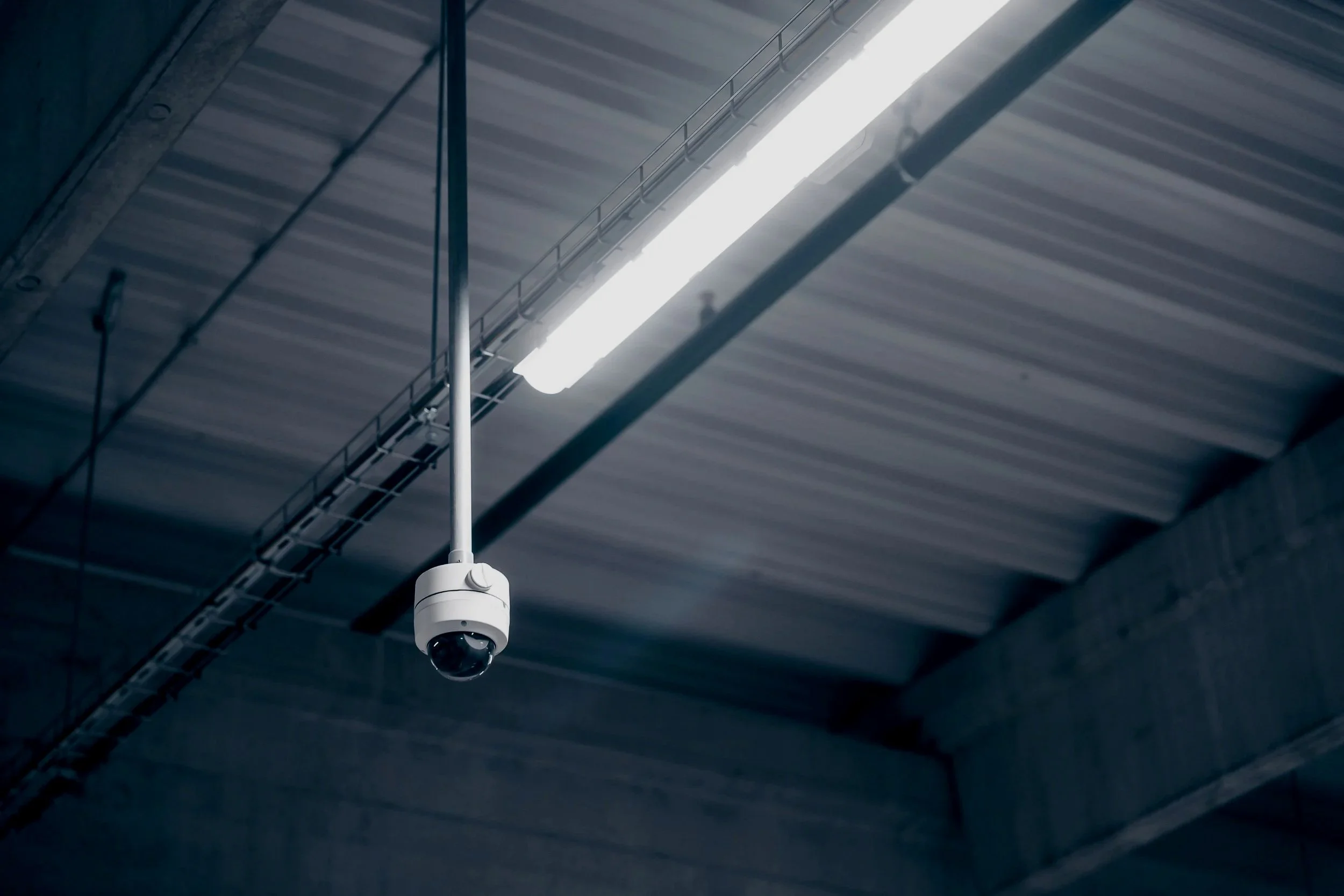 Security camera hanging from the ceiling of an industrial building with exposed metal roof panels and fluorescent lighting.