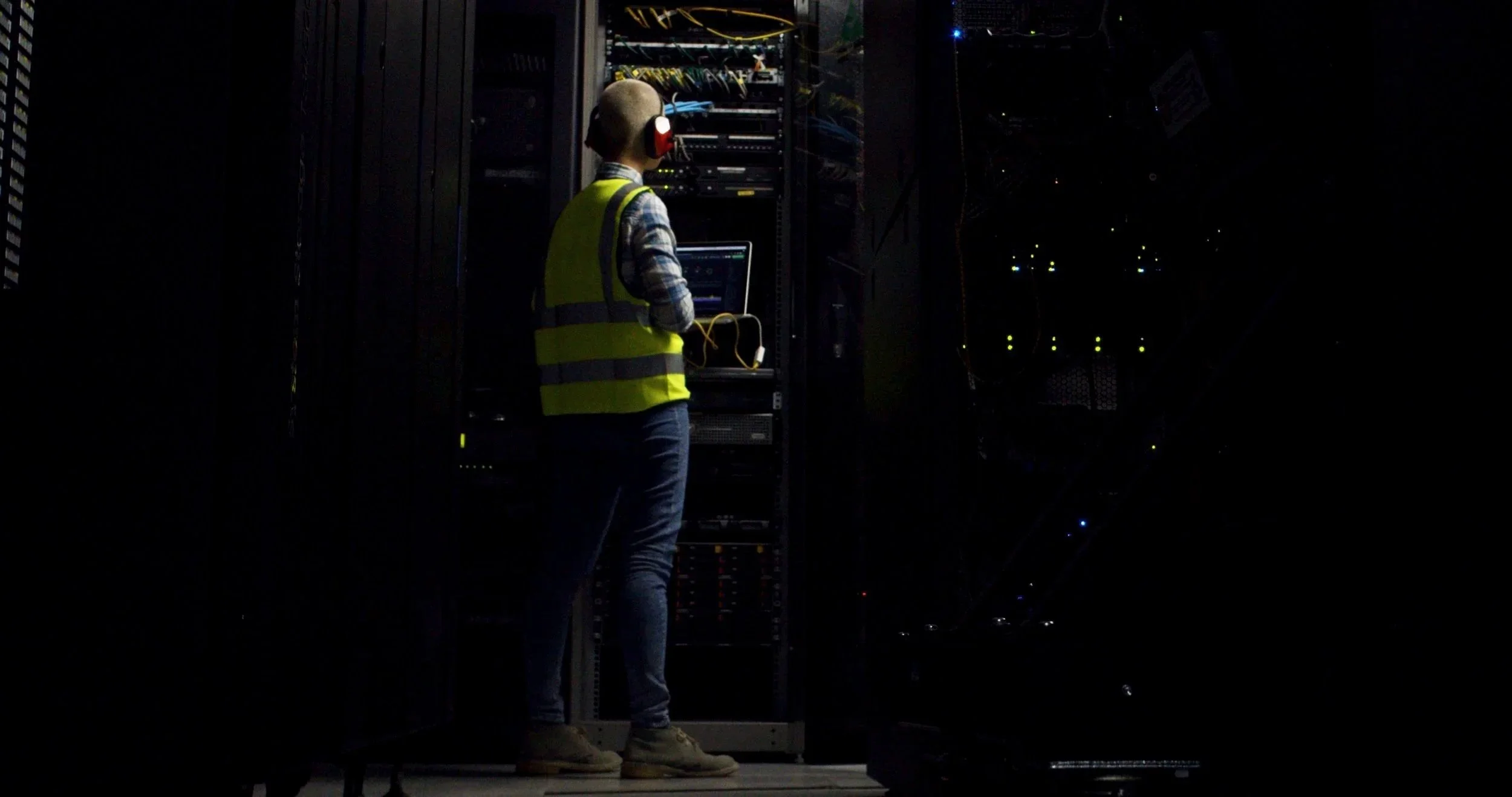 A person wearing a yellow safety vest, plaid shirt, and headphones working on a laptop in a dark data center or server room, surrounded by server racks with blinking lights.