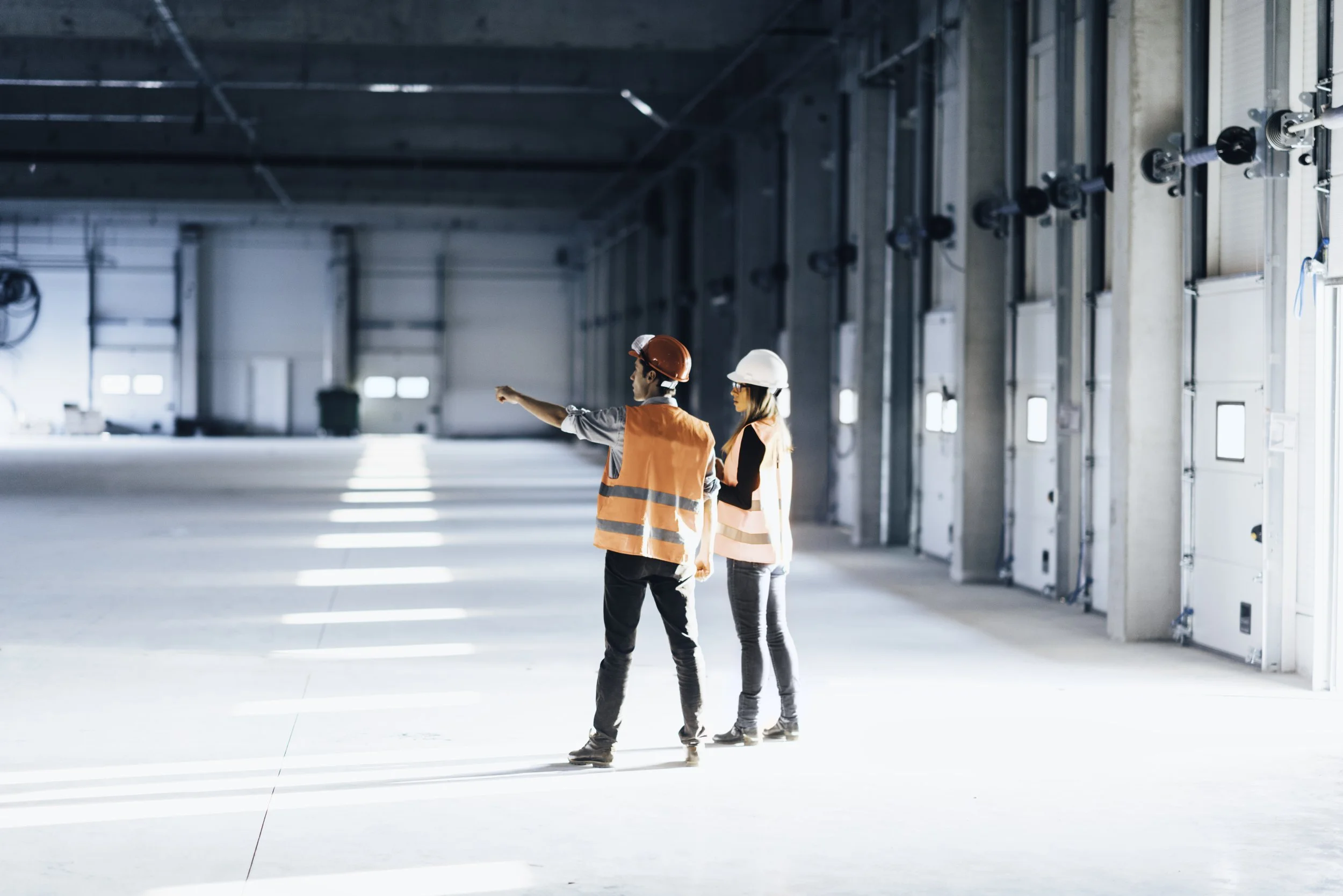 Two construction workers, a man and a woman, wearing safety helmets and vests, standing inside a large industrial building, with the man pointing towards something.